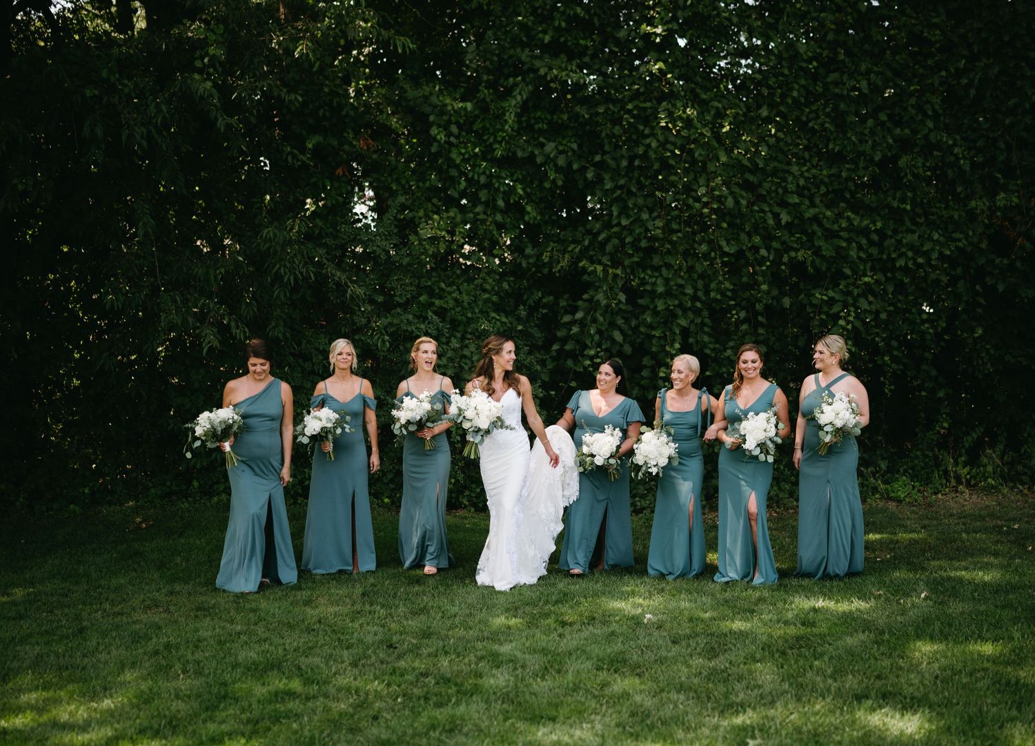 Wedding party in blue dresses and white attire pose together on grassy lawn during outdoor celebration.