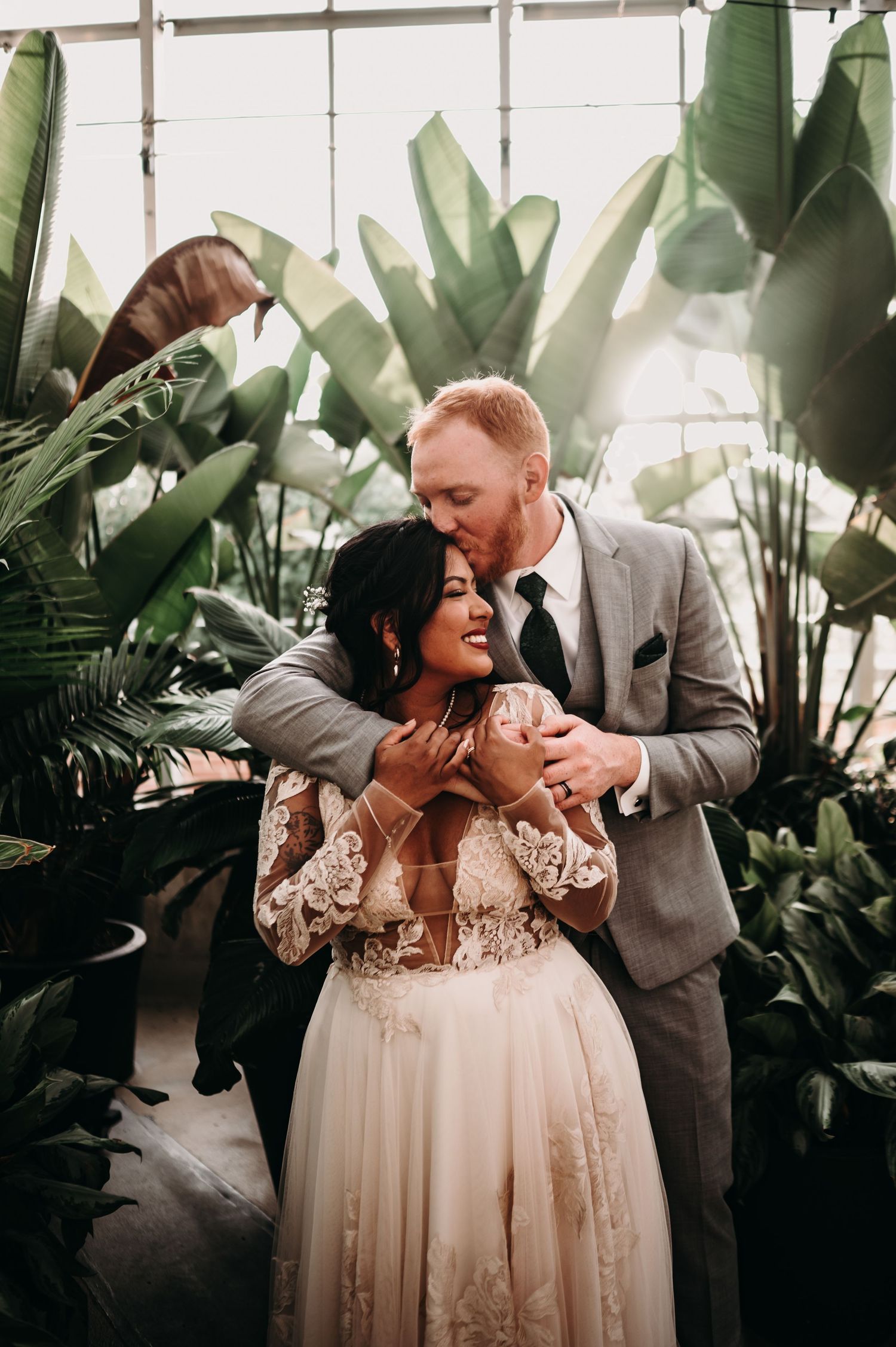 Intimate wedding portrait showing tender embrace amidst green plants in a greenhouse.