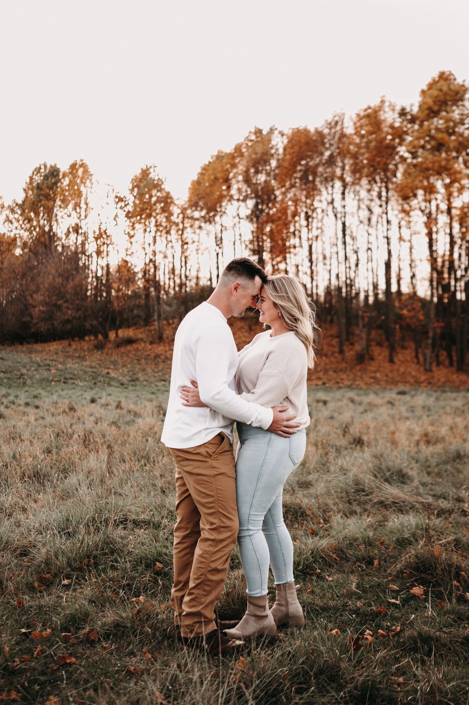 Romantic couple embracing in field with fall foliage and warm sunset lighting.