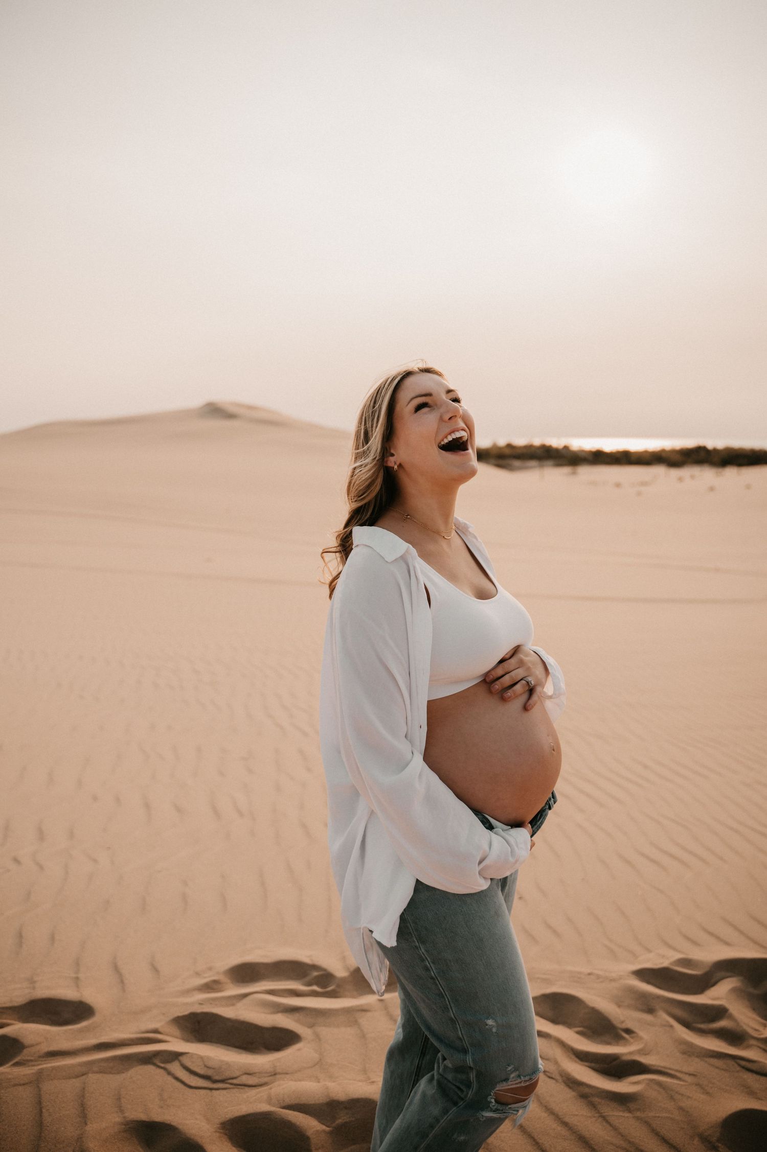 Joyful pregnant woman laughs while standing on desert sand dunes at sunset.