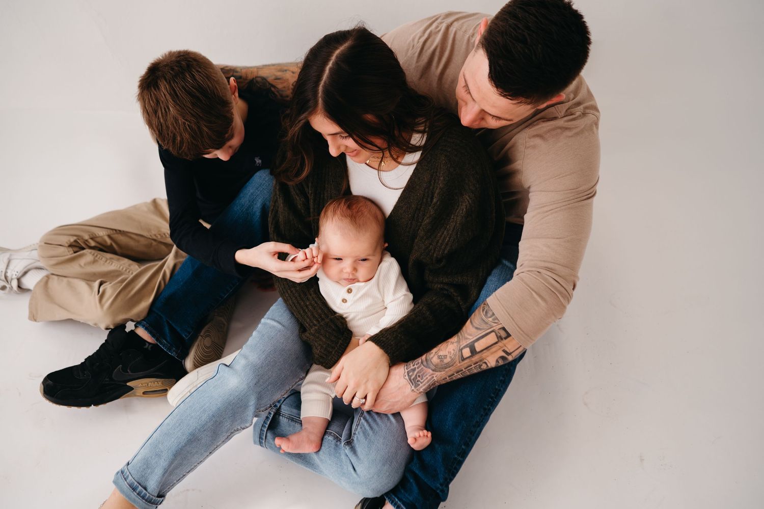 Family sits on floor together wearing casual clothing while interacting with young infant in cozy indoor setting.