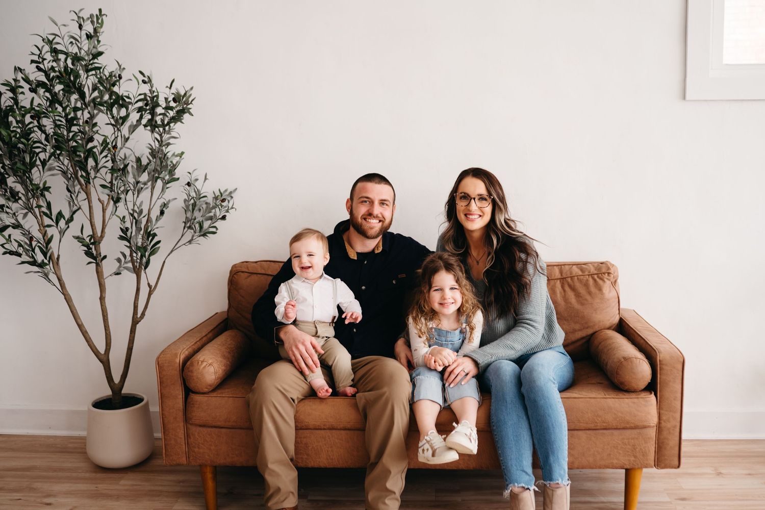 A family of four sits together on a brown leather couch next to a small indoor olive tree in a bright living room.