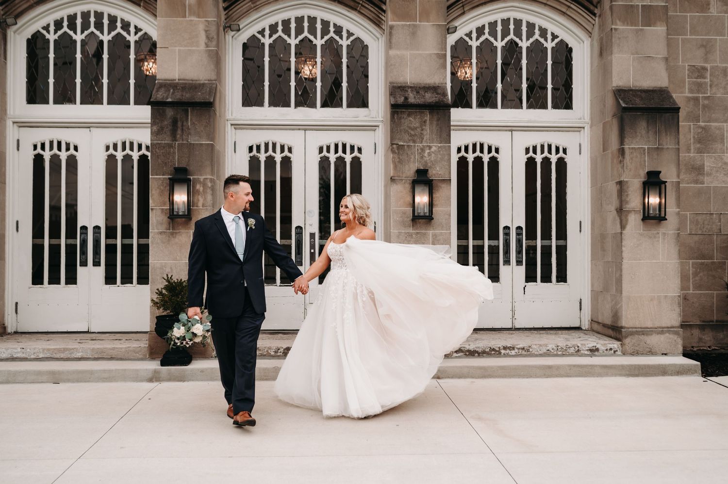 A couple walks together outside a Gothic-style building with arched windows.