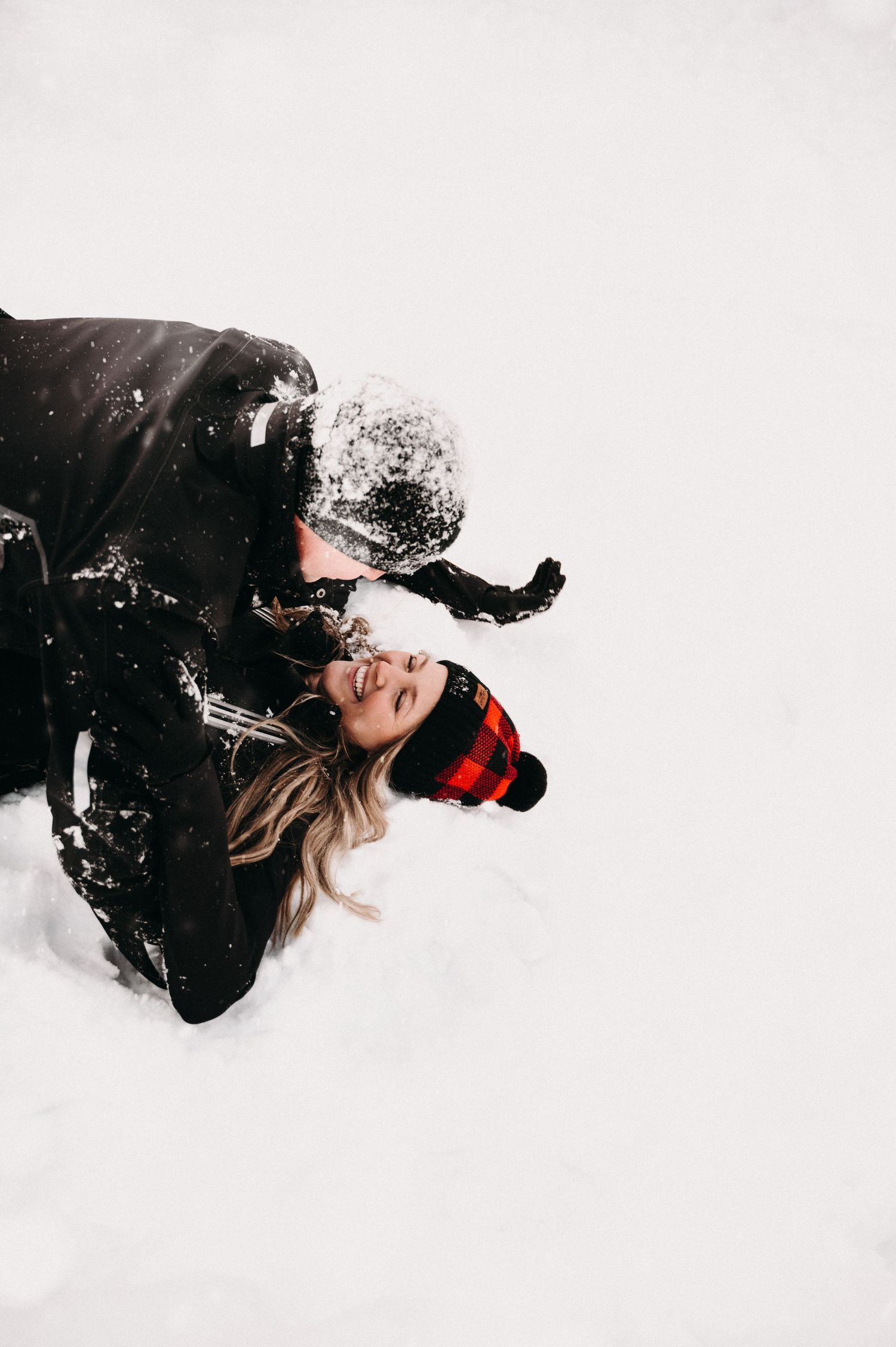 Two people in black winter coats playing in the snow with red plaid hats.