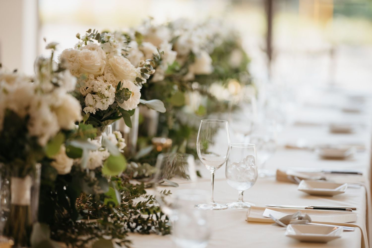 Elegant wedding reception tables decorated with white floral centerpieces and place settings in a light-filled venue.