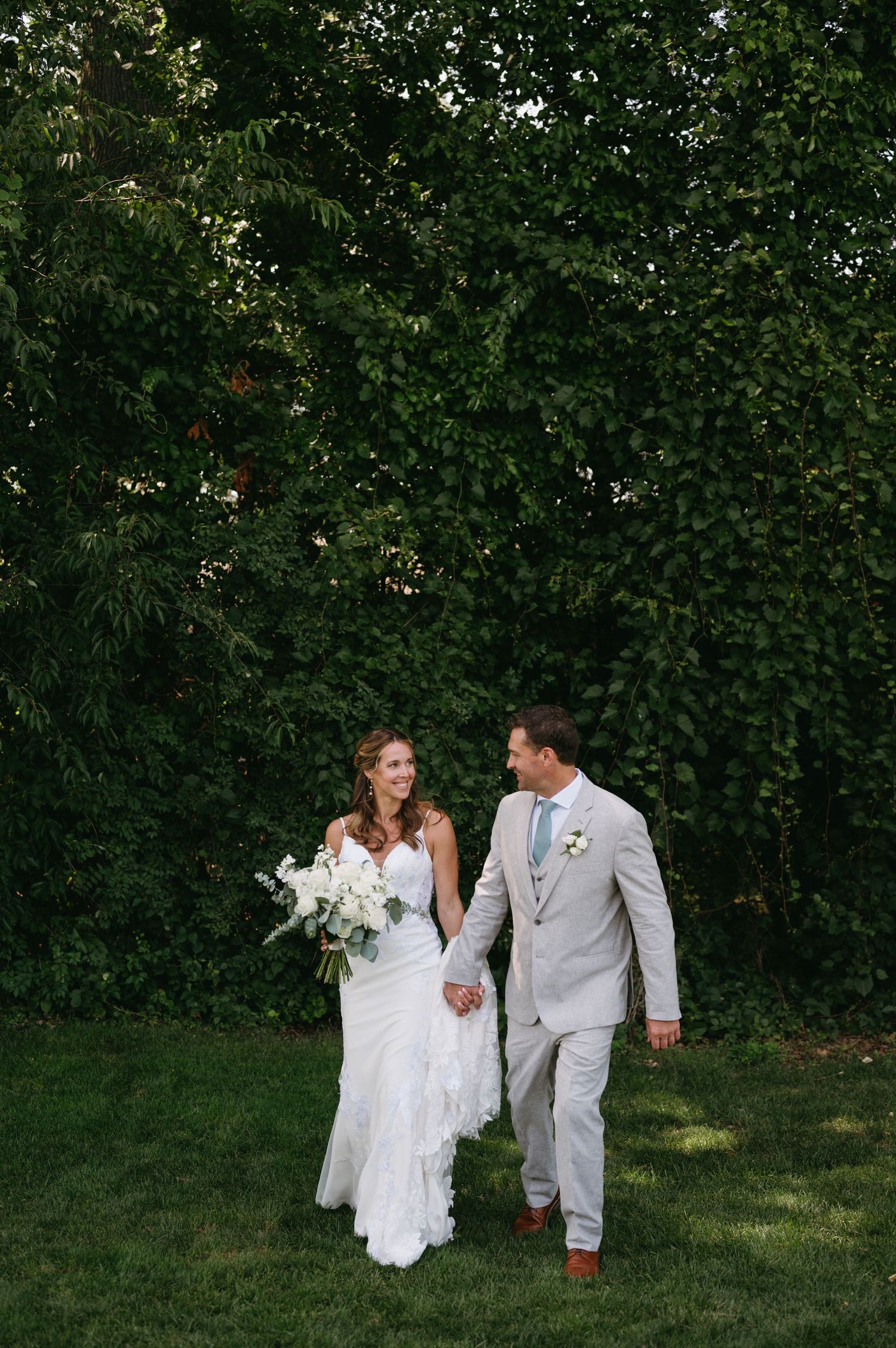 A couple in wedding attire walks together on a grassy lawn surrounded by lush green foliage.