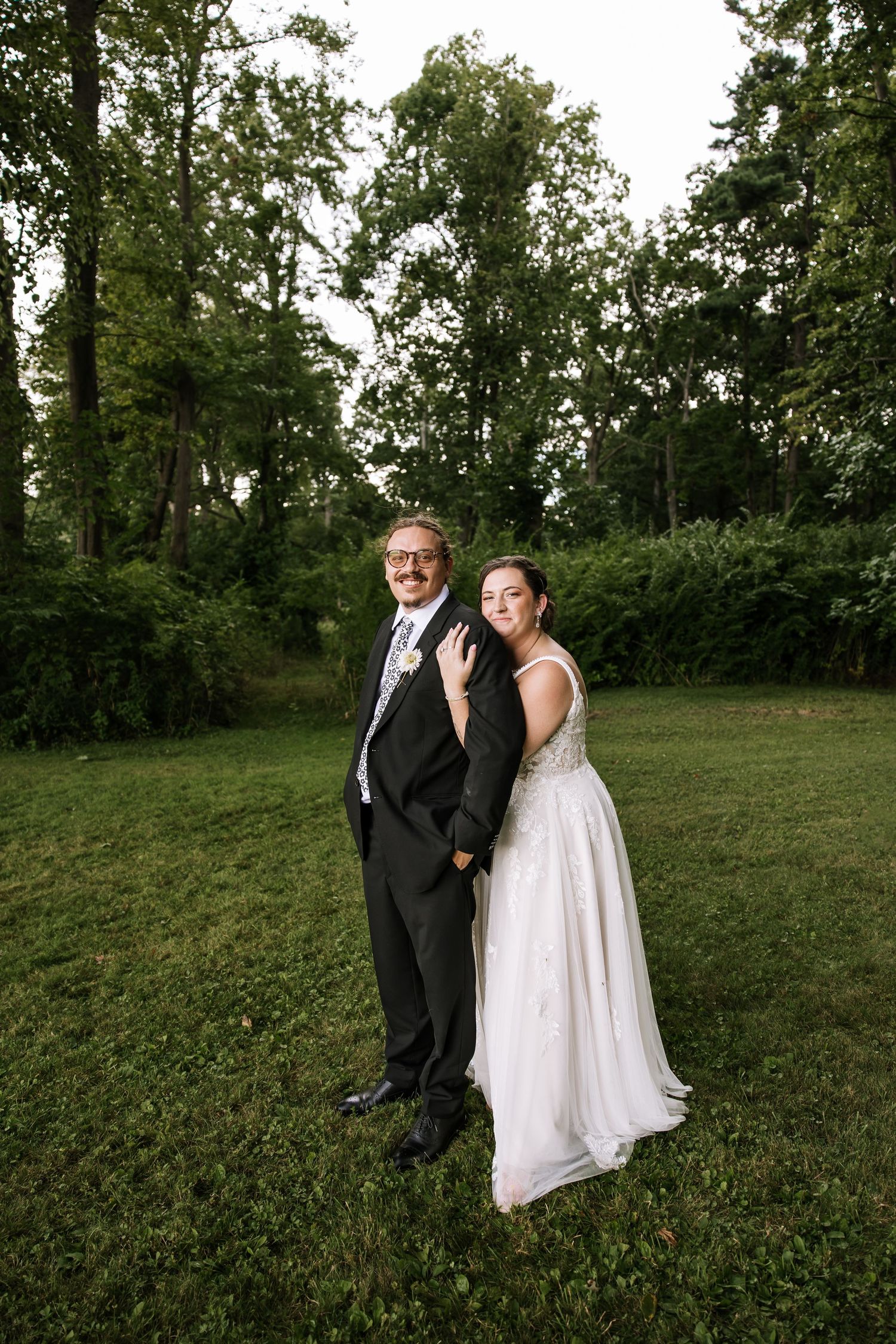 A couple in wedding attire poses together on a grassy lawn surrounded by trees during an outdoor photo session.