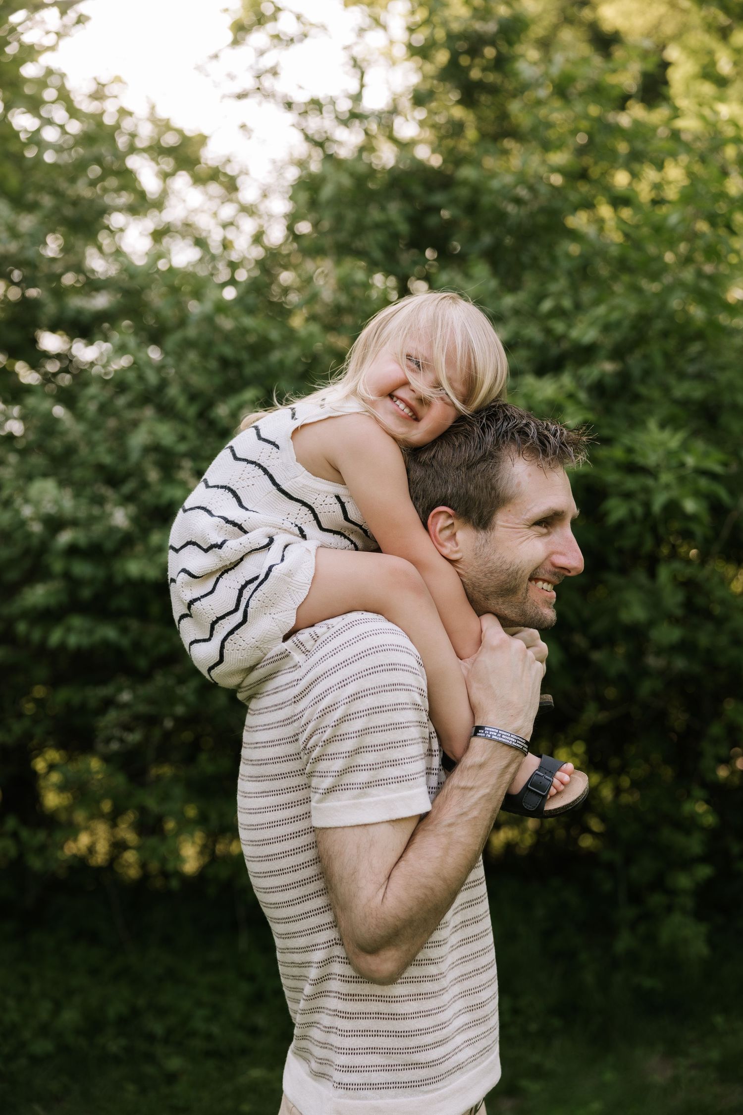 A smiling father carries his laughing blonde child on his shoulders outdoors among green trees.
