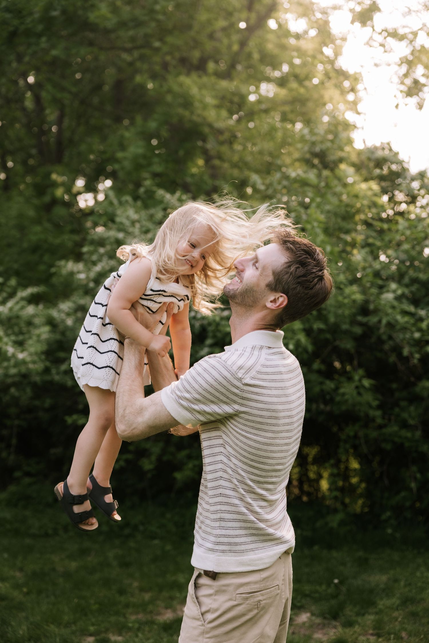 A smiling father lifts his joyful blonde toddler daughter into the air in a sunlit backyard.