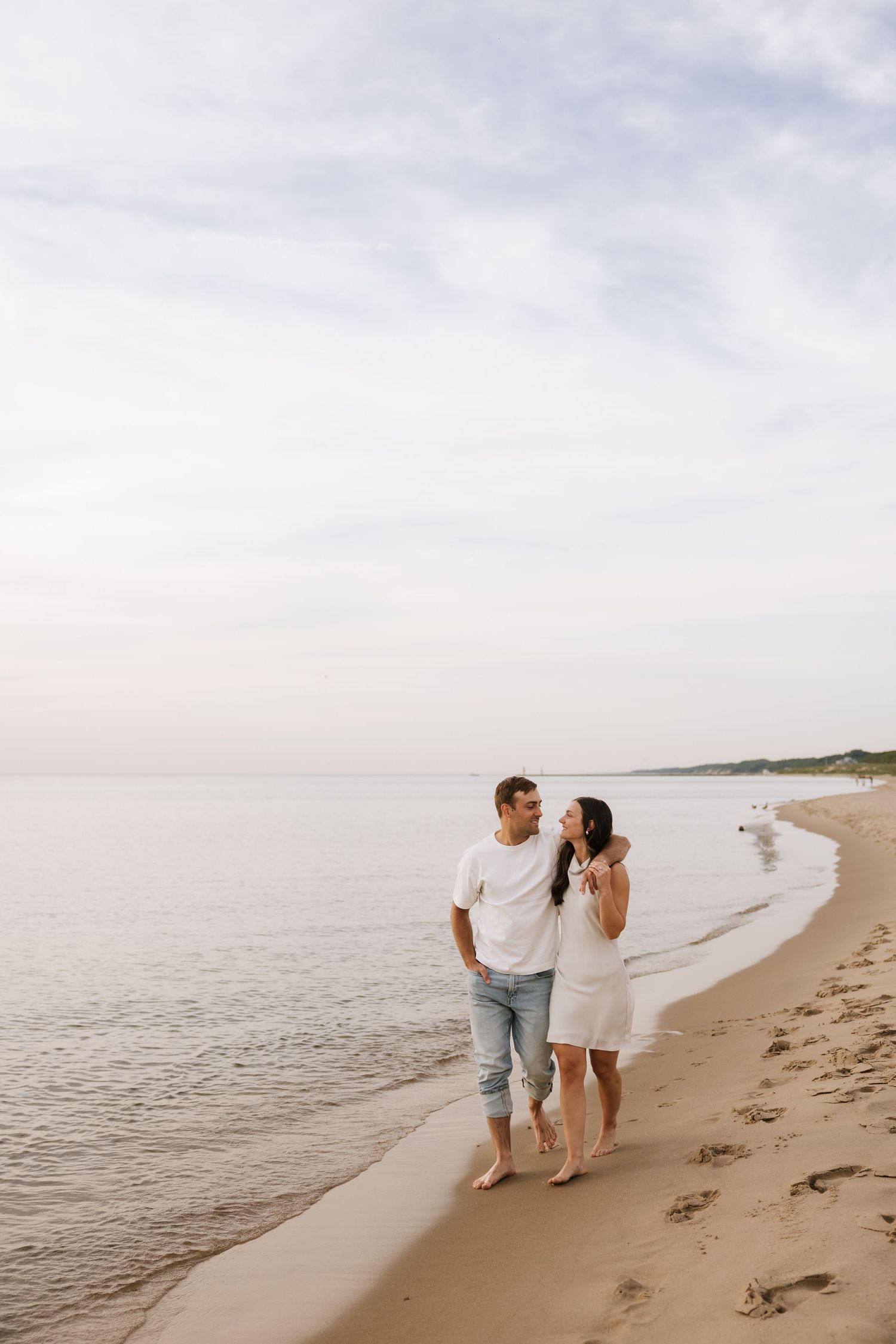 Couple walking barefoot along shoreline holding hands, wearing white outfits on a calm overcast beach day.