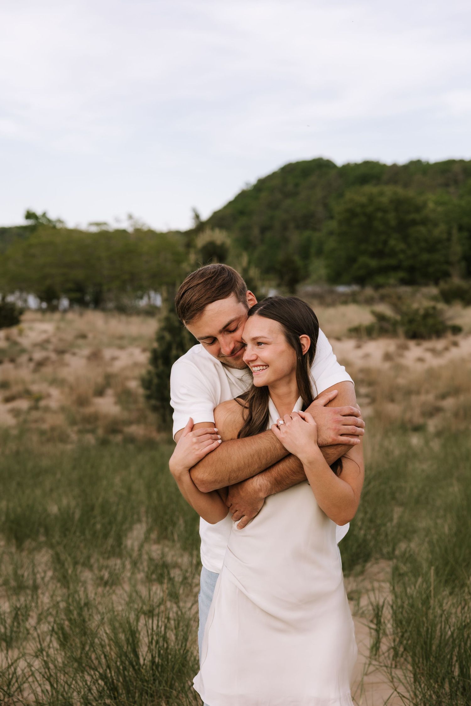 Man in white shirt hugging woman in white dress from behind, both smiling in grassy outdoor setting.