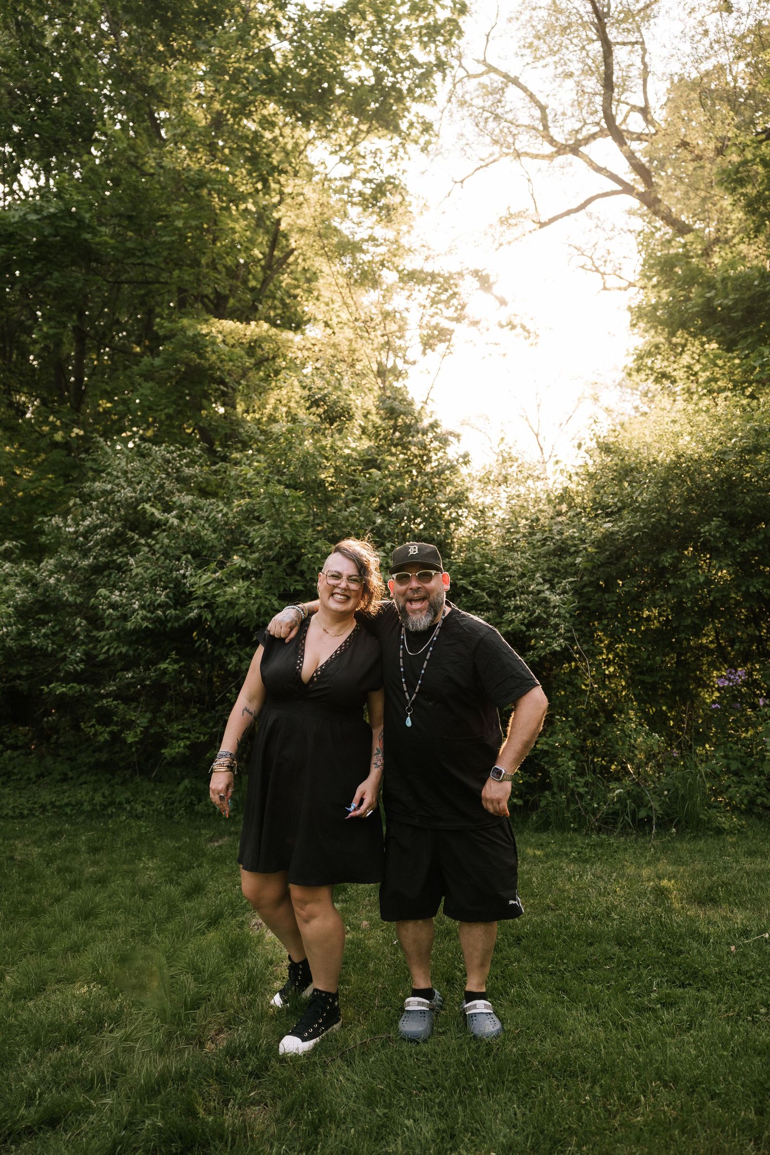 Smiling couple in matching black outfits posing together on green grass surrounded by lush trees in golden hour light.