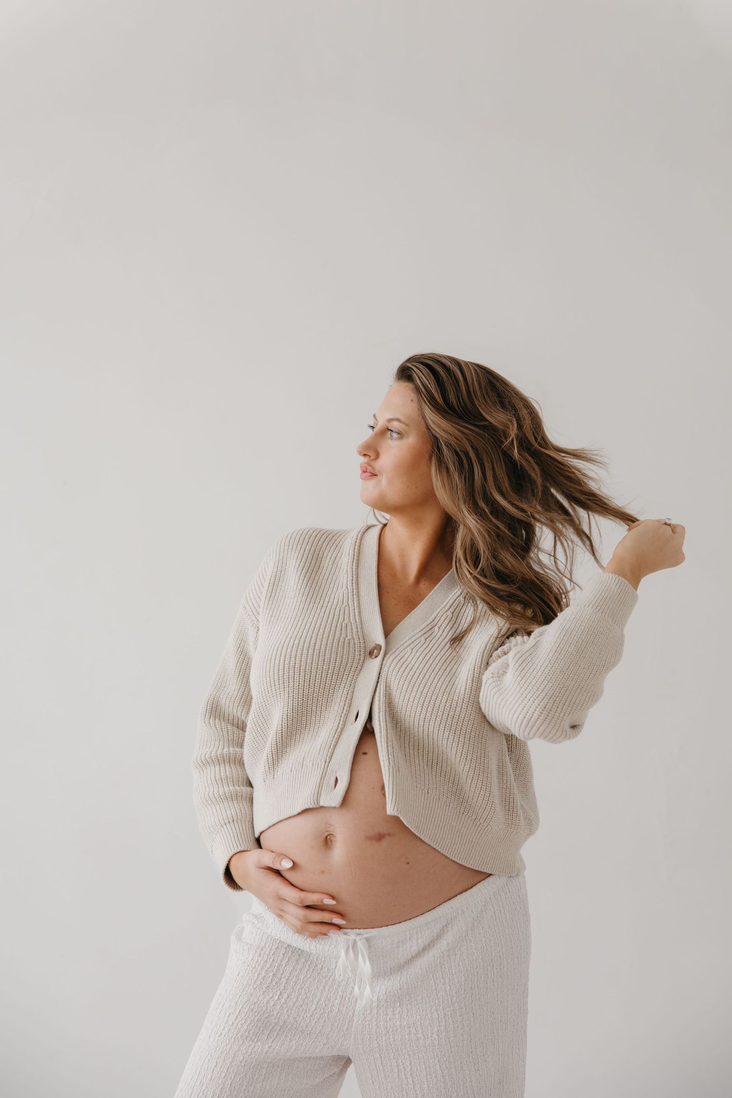 Pregnant woman in beige knit cardigan and white pants cradles her baby bump while posing against a white background.