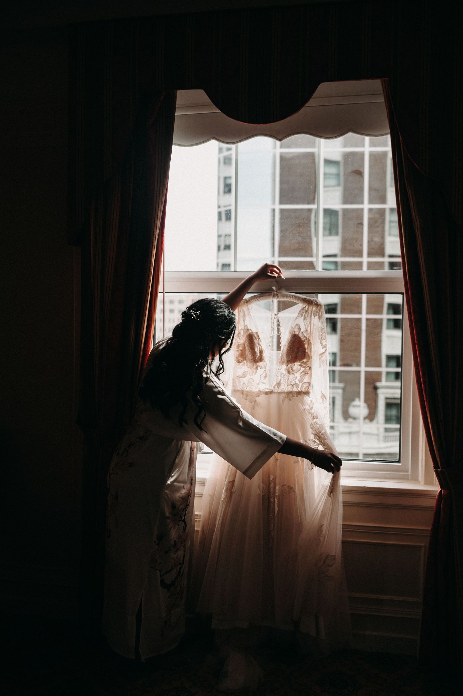 A wedding dress hangs in silhouette by a window as someone adjusts it in warm natural light.