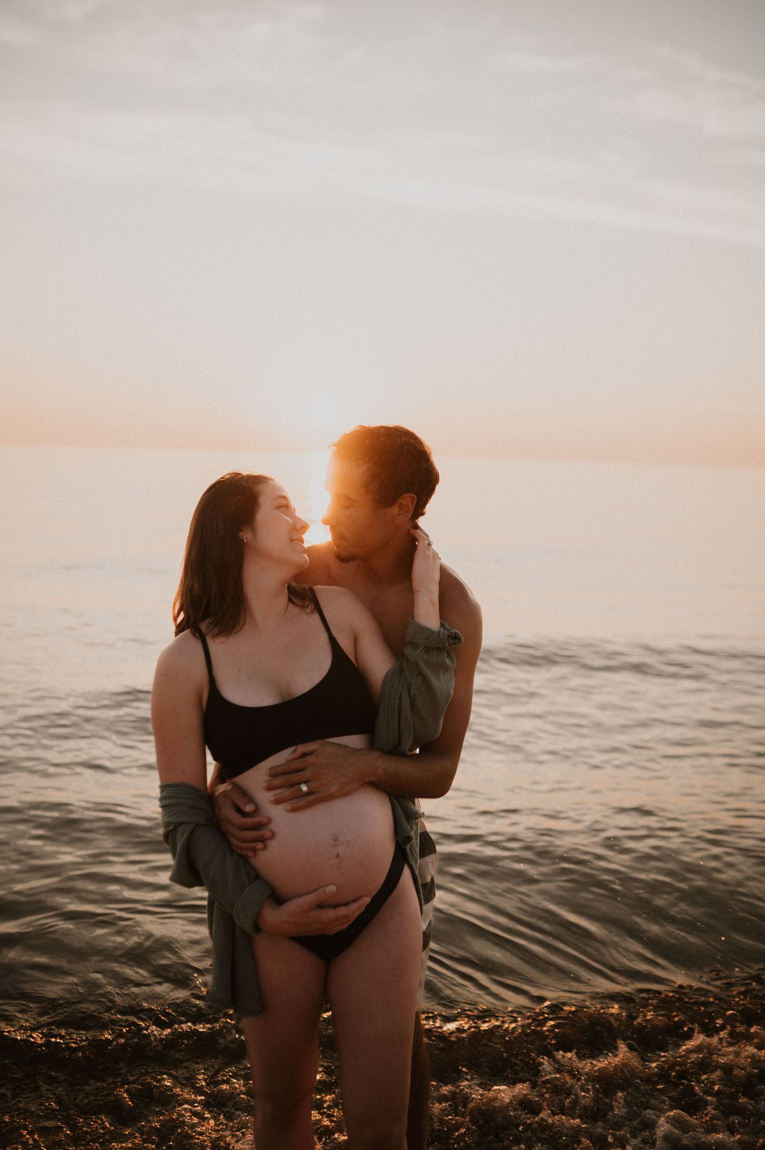 Tender embrace between expectant couple during sunset beach photoshoot.