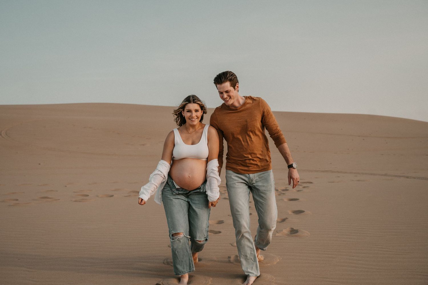 A couple walks together on desert sand dunes during a maternity photoshoot.