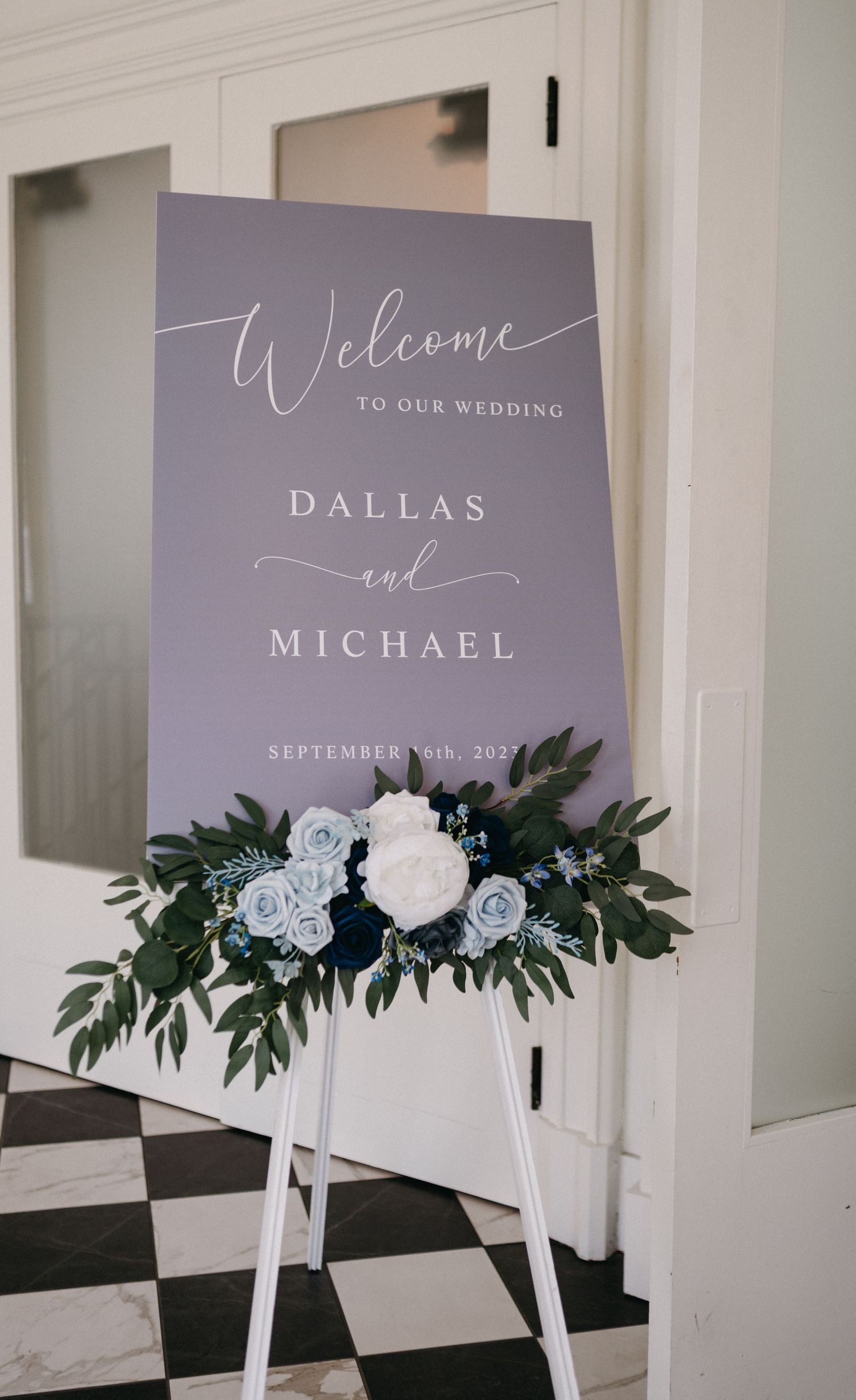 Elegant grey wedding welcome sign with white text and floral arrangement at the bottom.