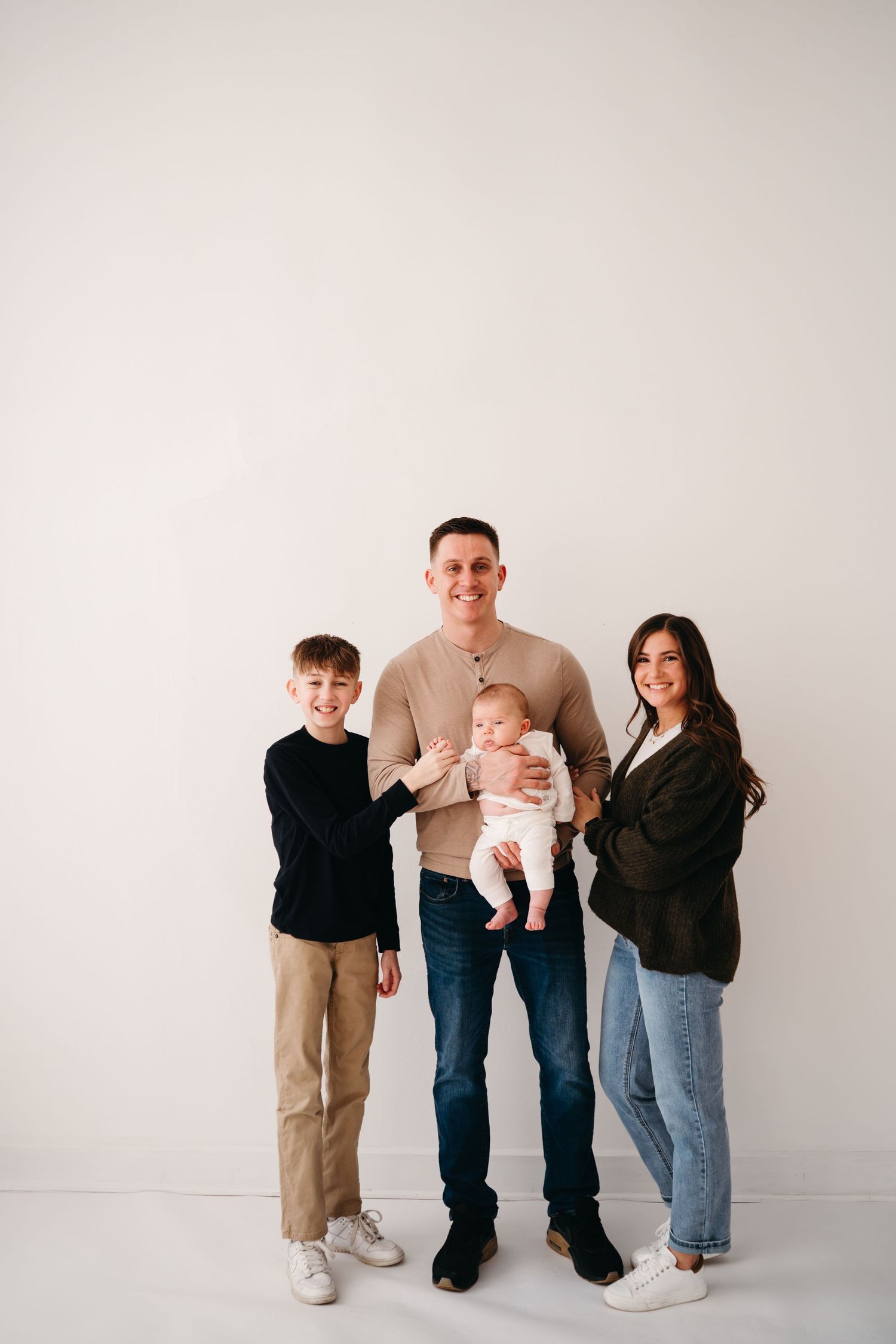 A family of four stands together against a white wall wearing casual clothing in a minimalist studio setting.