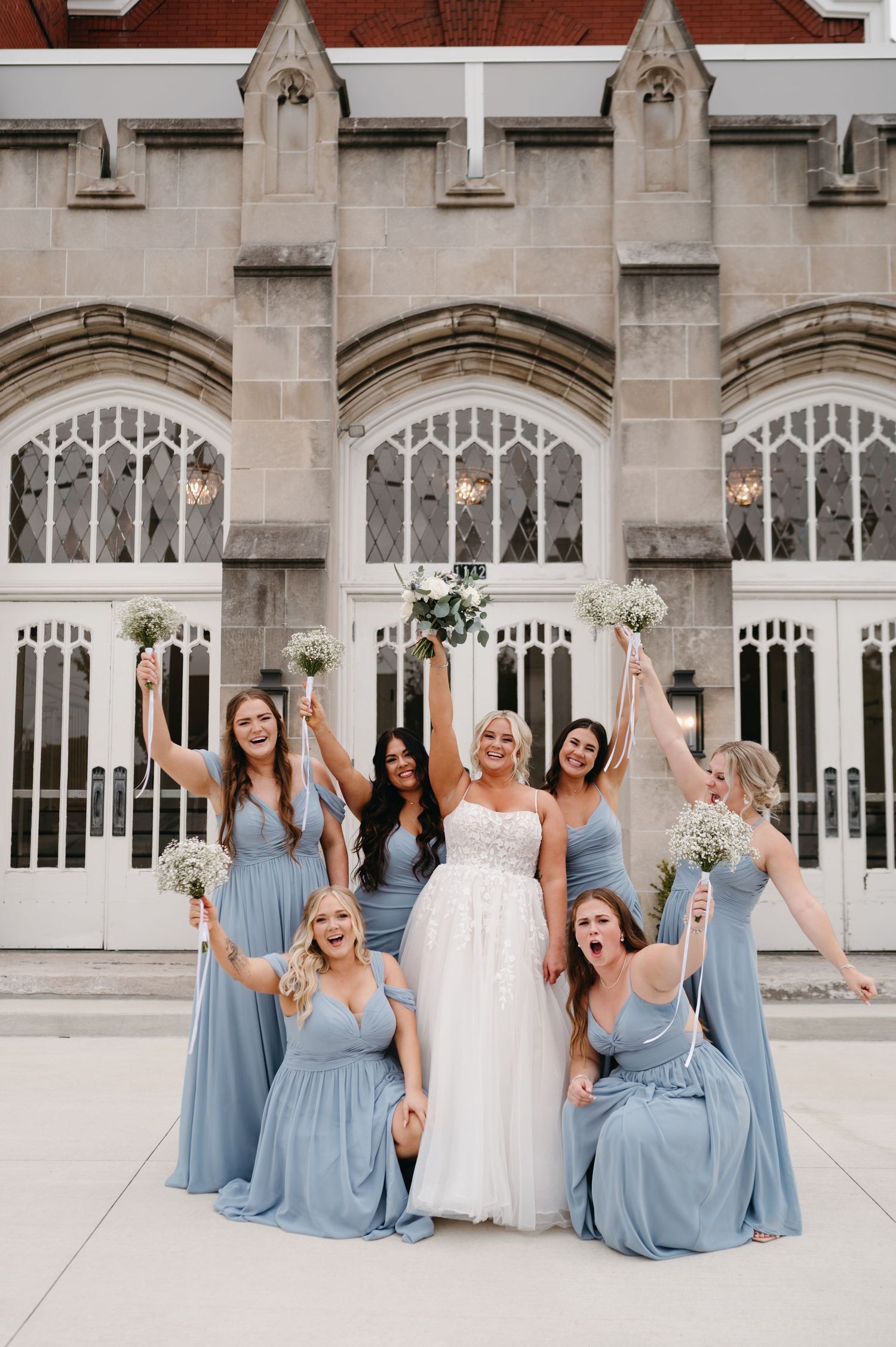 Celebratory bridal party in blue dresses cheers excitedly in front of church windows.