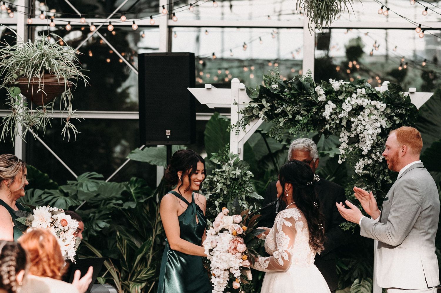 Wedding party stands before lush greenery backdrop during ceremony at indoor botanical venue.