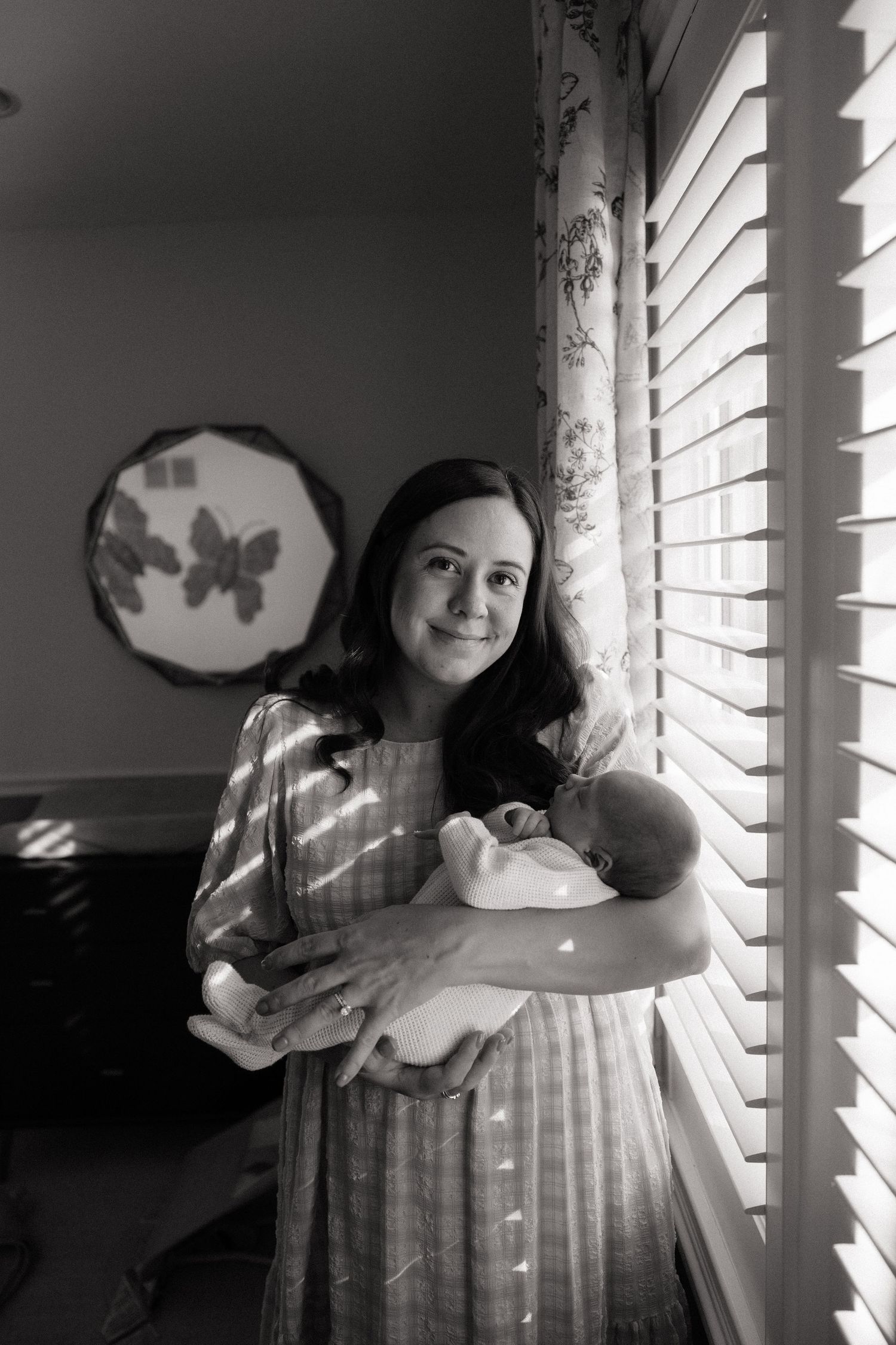 Black and white portrait of mother and newborn by window with filtered light through blinds.