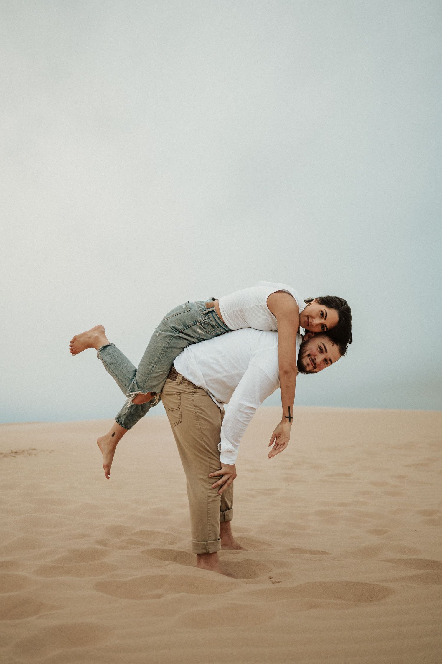 A playful couple poses on sand dunes with one person piggybacking on the other.