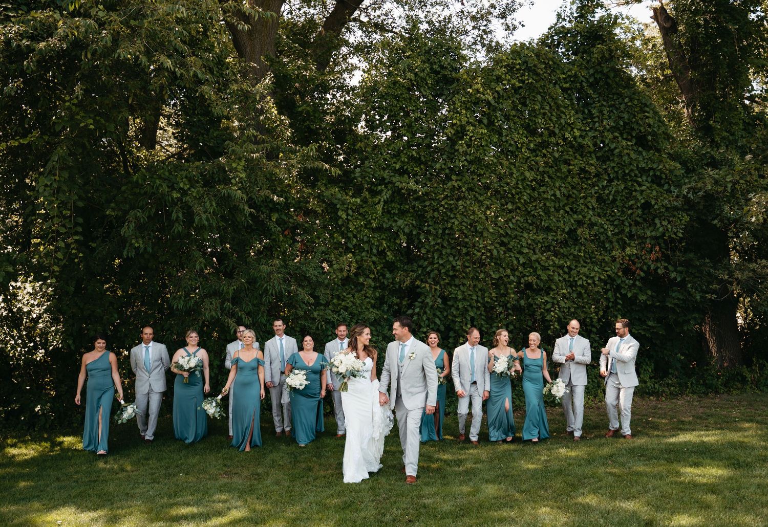 Wedding party members lined up in formal attire against a lush green outdoor backdrop at sunset.