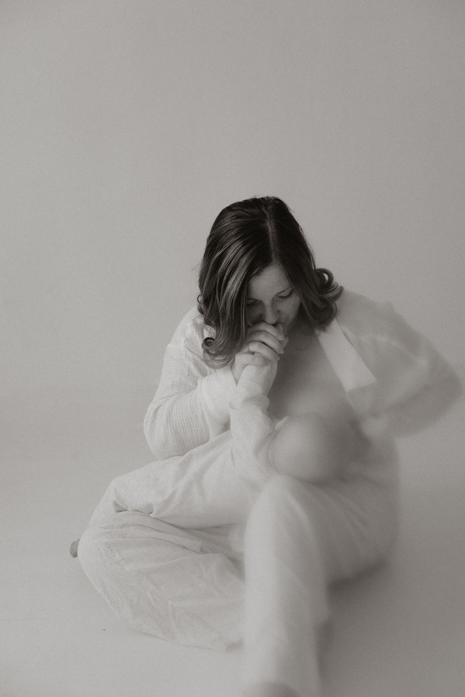A pregnant woman in a flowing white dress sits gracefully, head bowed, in a serene black and white maternity portrait.