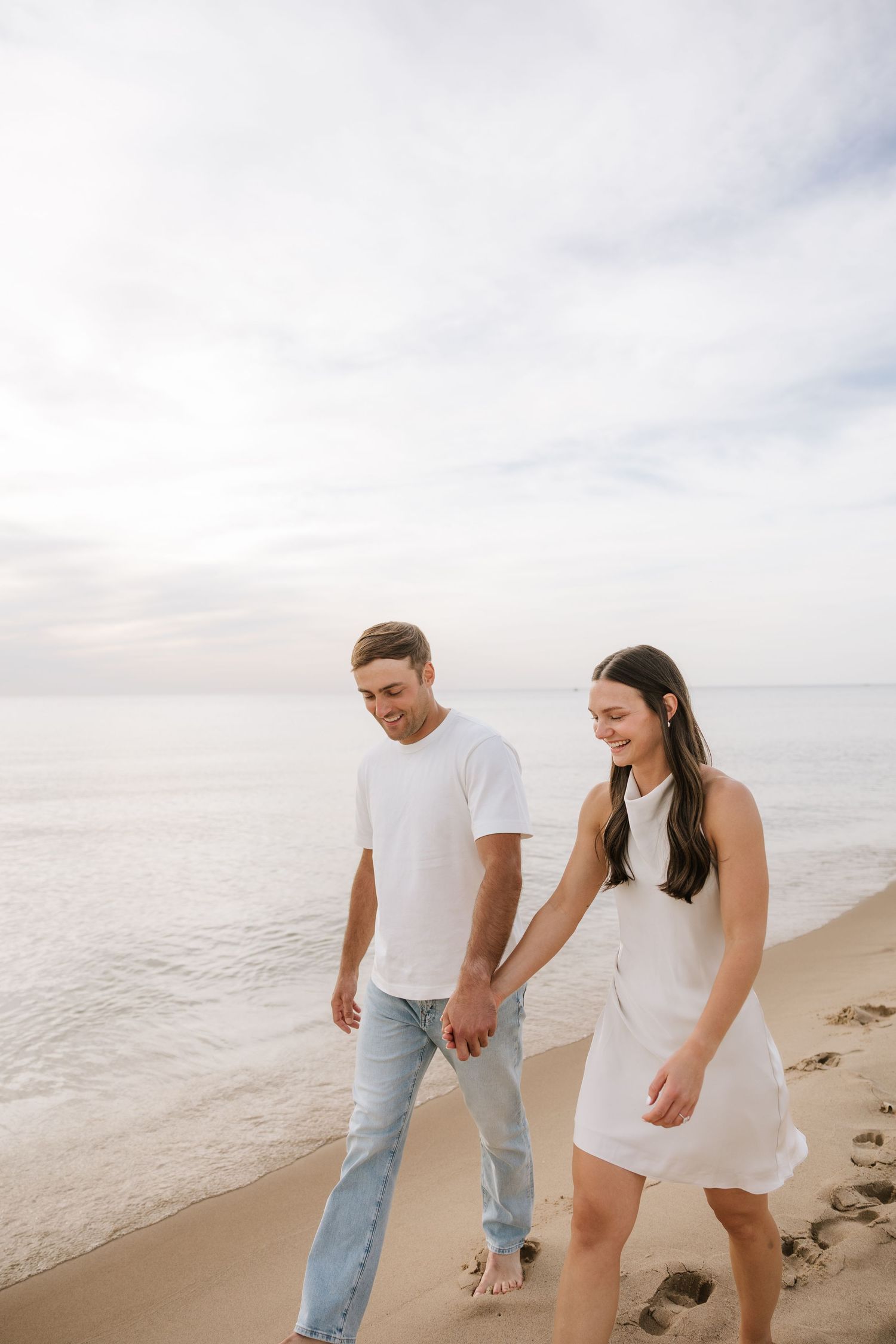 Smiling couple holding hands and walking barefoot along a sandy beach with calm ocean waves behind them.