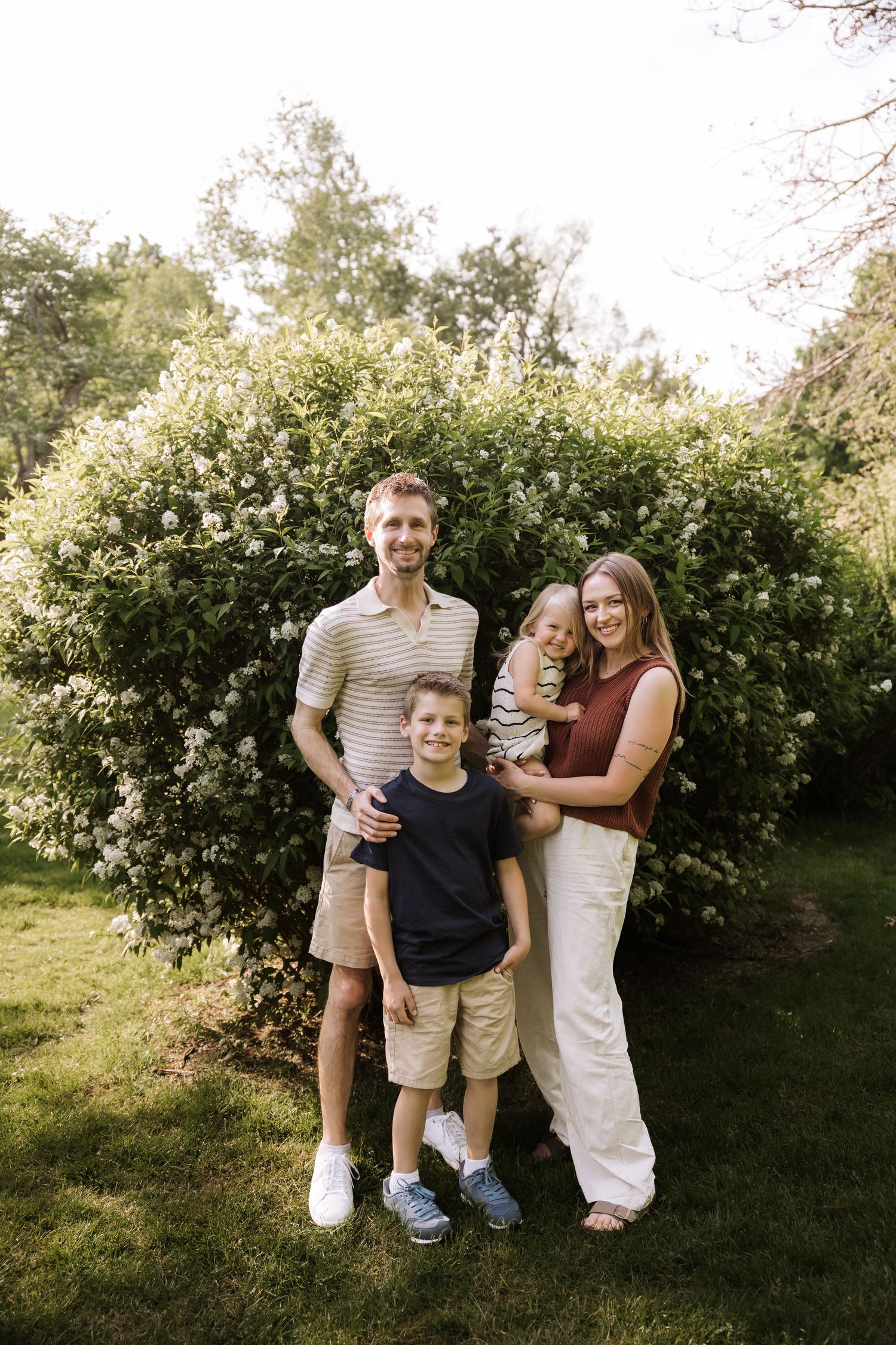 Happy family of four posing outdoors in front of lush flowering bushes on a sunny day.