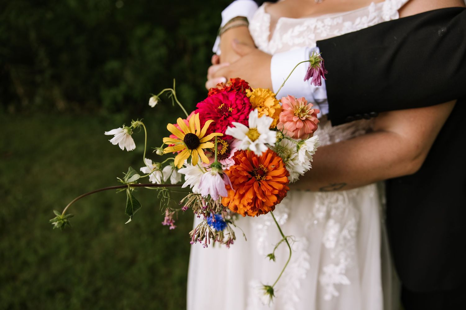 A close-up view of a colorful bridal bouquet with sunflowers, daisies and red flowers against a white wedding dress.