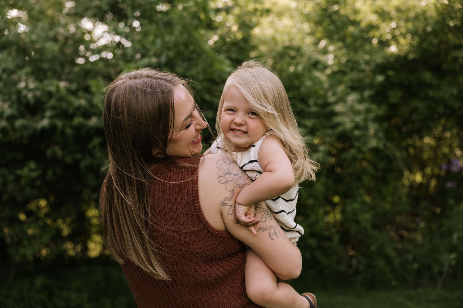 Mother holding her smiling blonde toddler daughter outdoors in green garden.