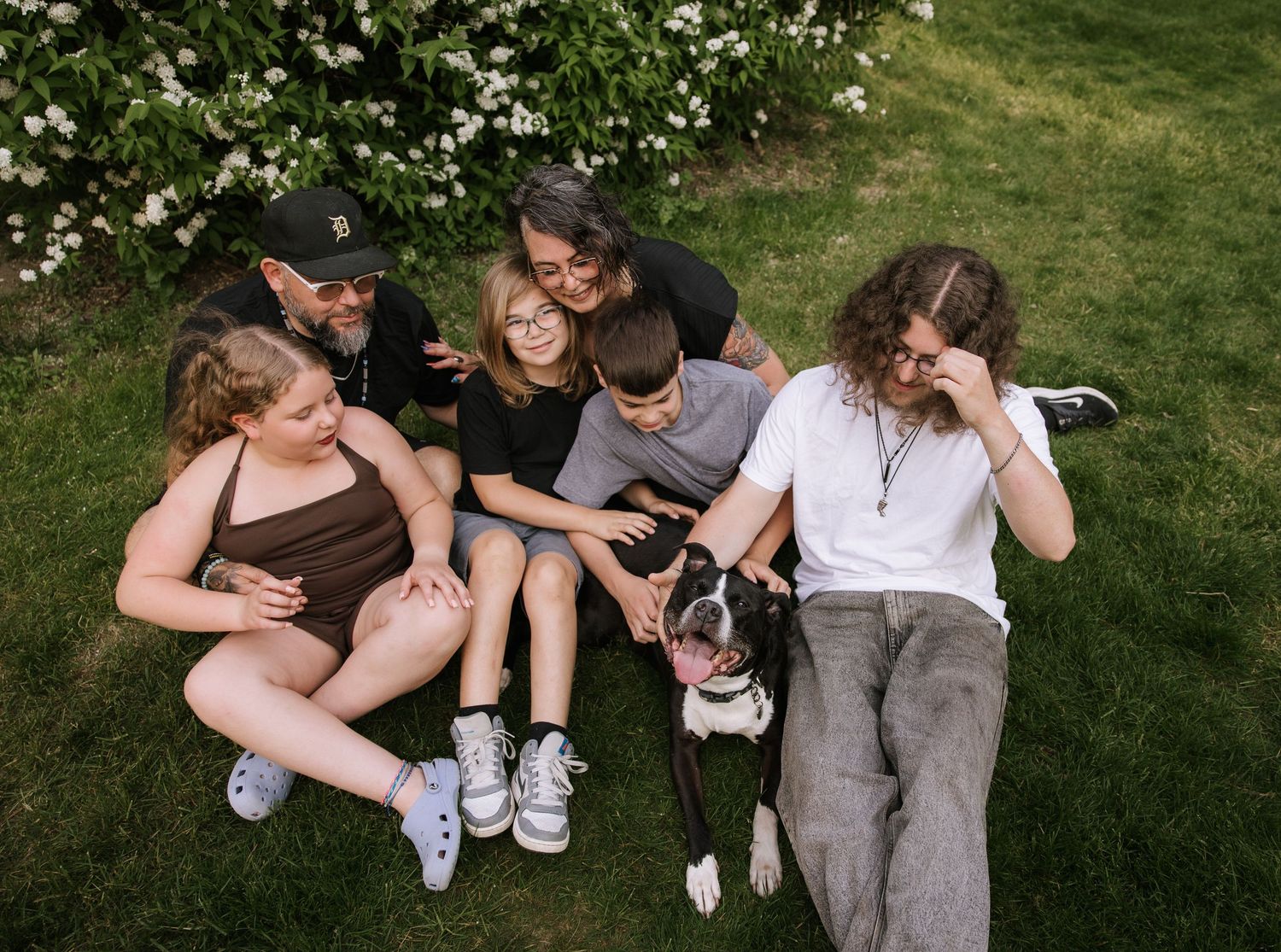 Family of five sitting on green grass together with their black and white pit bull dog.
