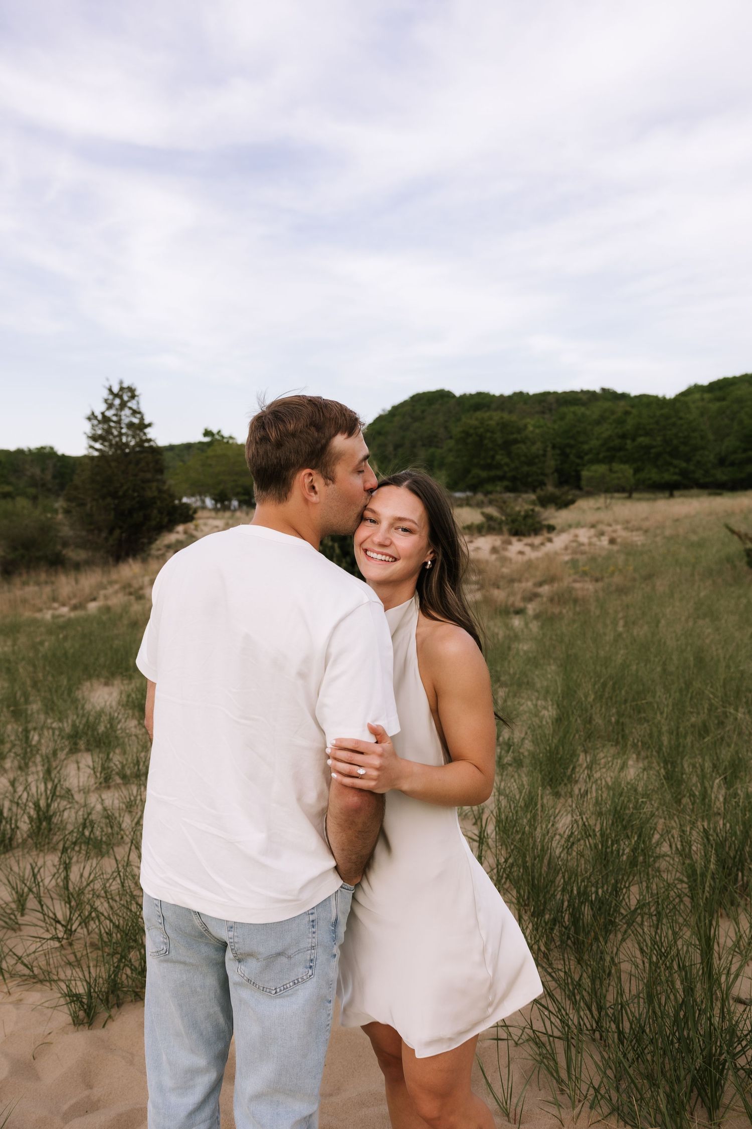 Happy couple dancing on beach sand dunes, man kissing smiling woman in white dress, lush green trees behind.