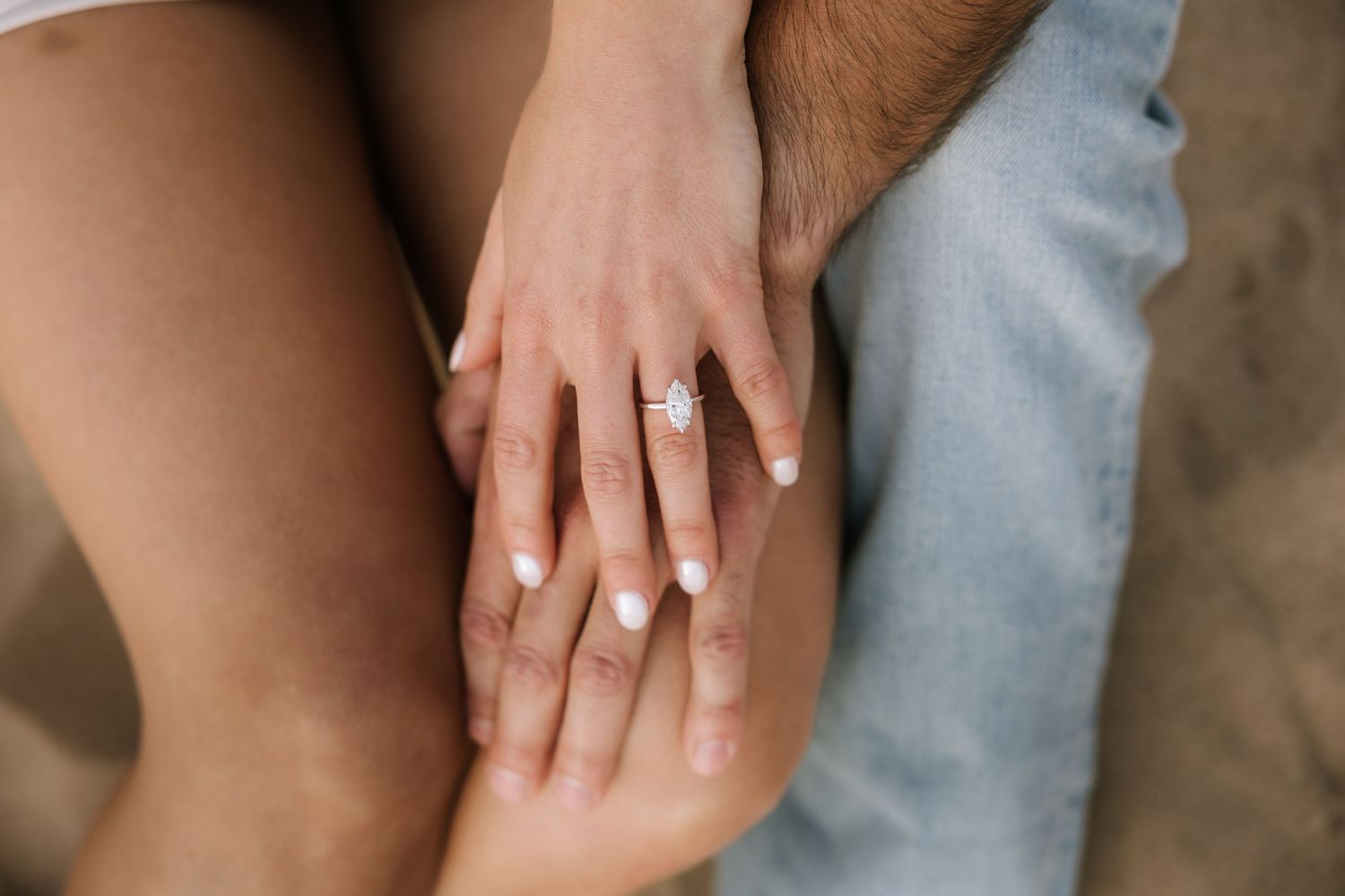 Close-up of an engagement ring with an oval diamond on a woman's hand resting on her partner's knee.