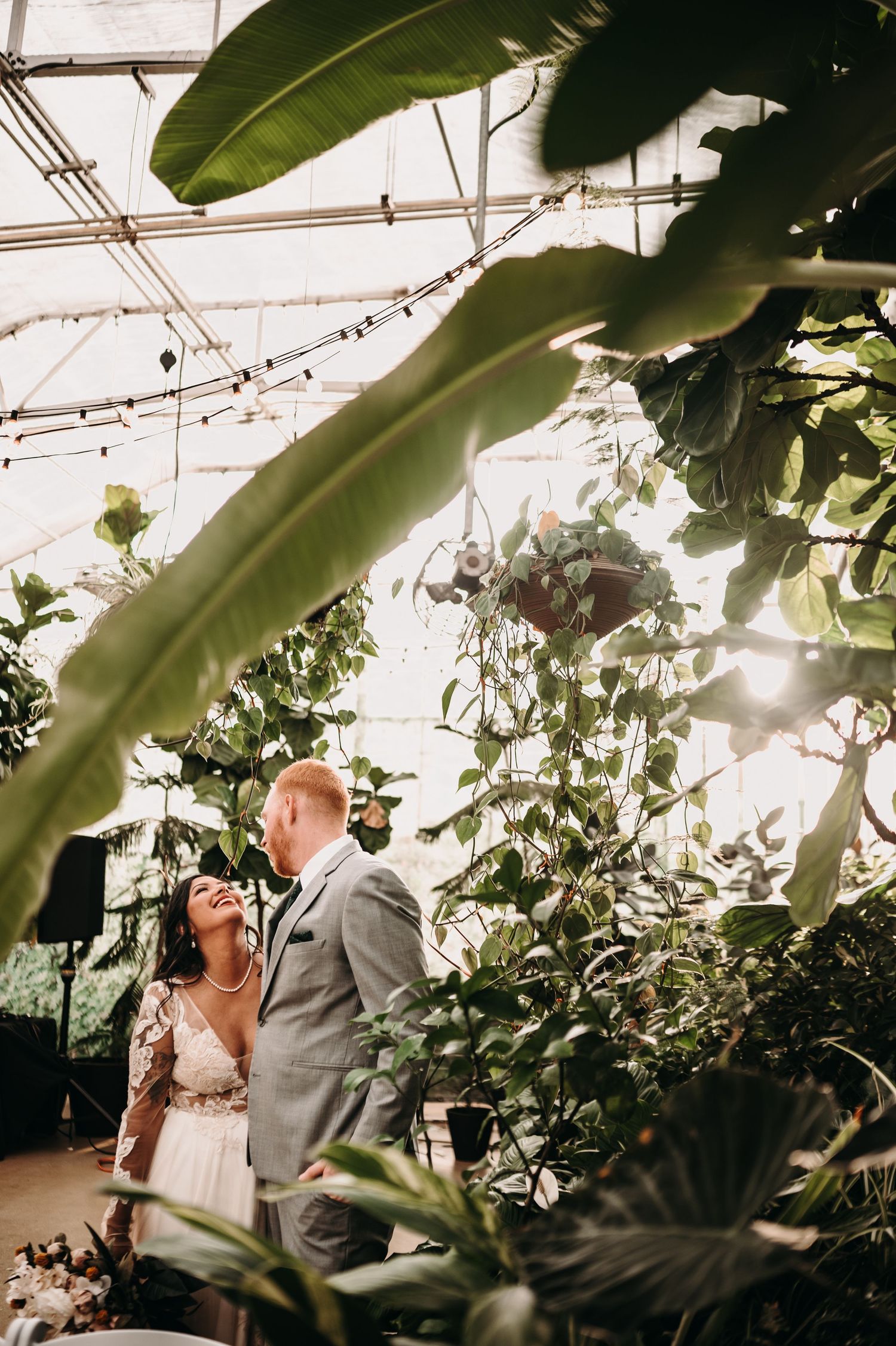 Sunlit greenhouse wedding portrait with tropical foliage and string lights overhead.