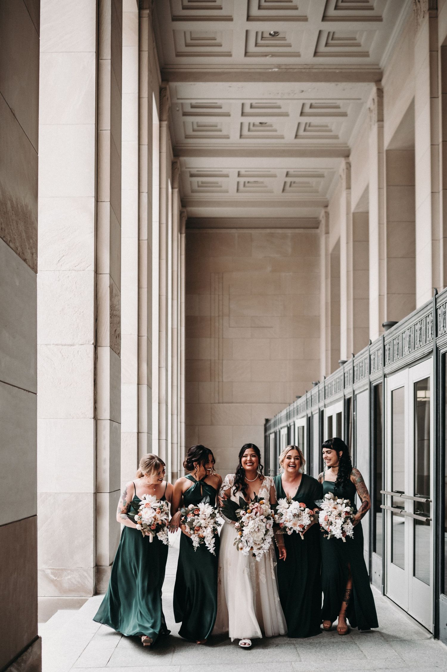 Wedding party in emerald green dresses poses elegantly in a columned hallway with floral bouquets.