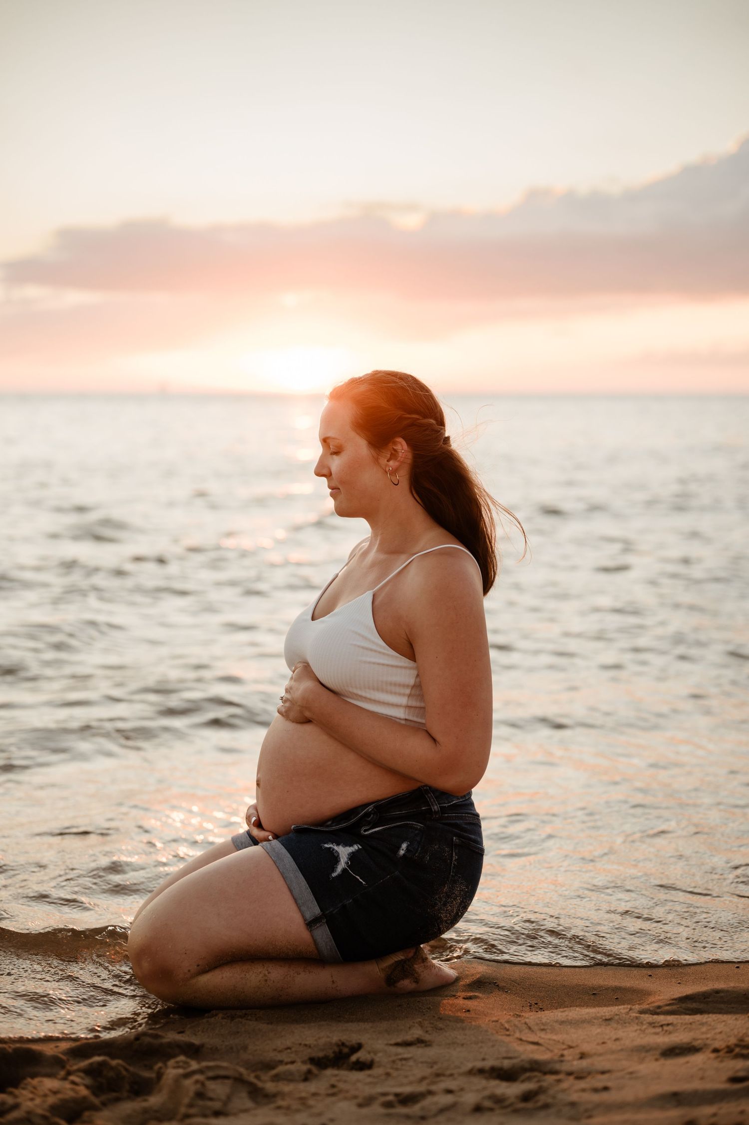 A pregnant woman sits on a sandy beach at sunset while waves lap at the shore in warm golden light.
