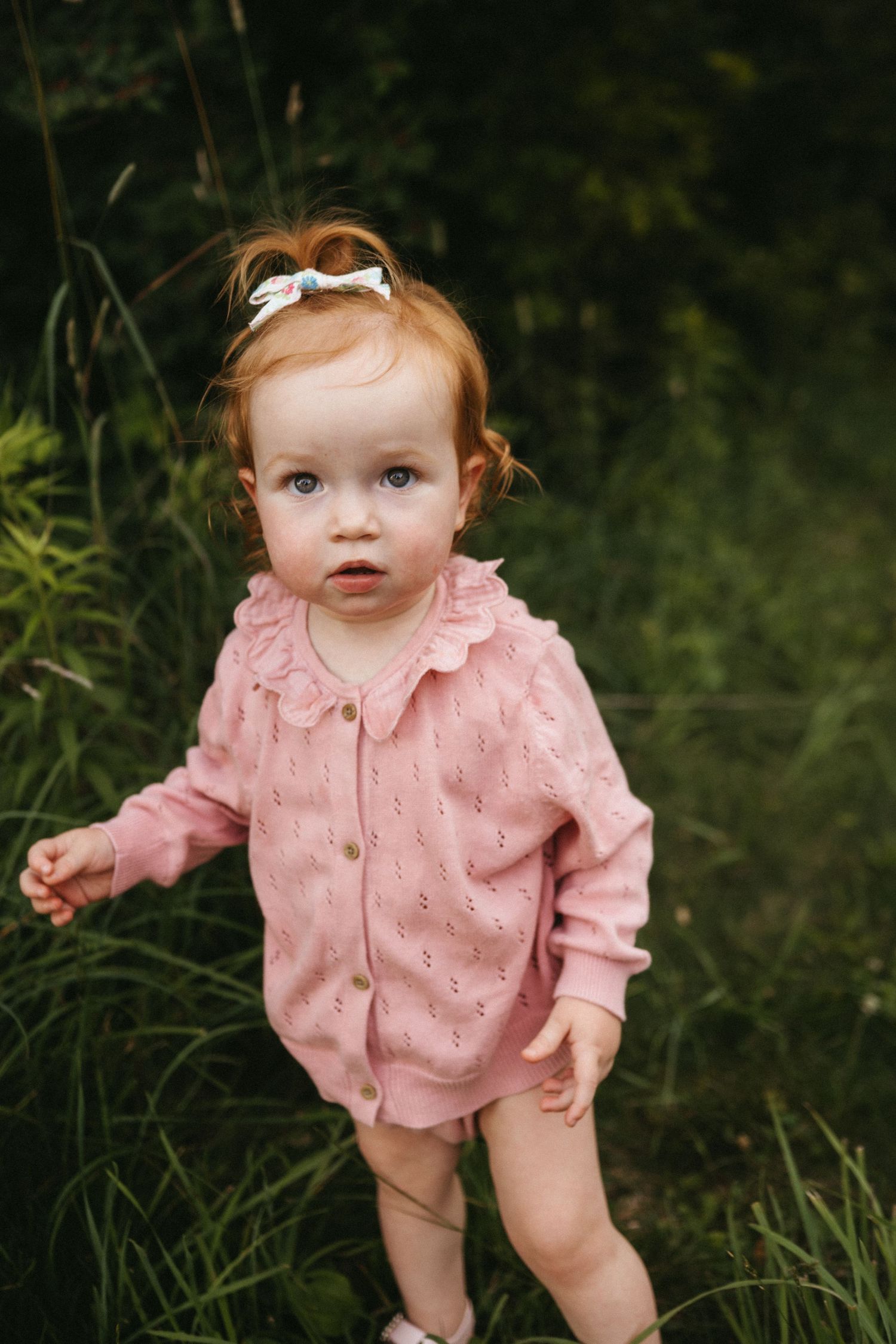 Baby wearing pink eyelet romper and white bow stands in tall grass outdoors.