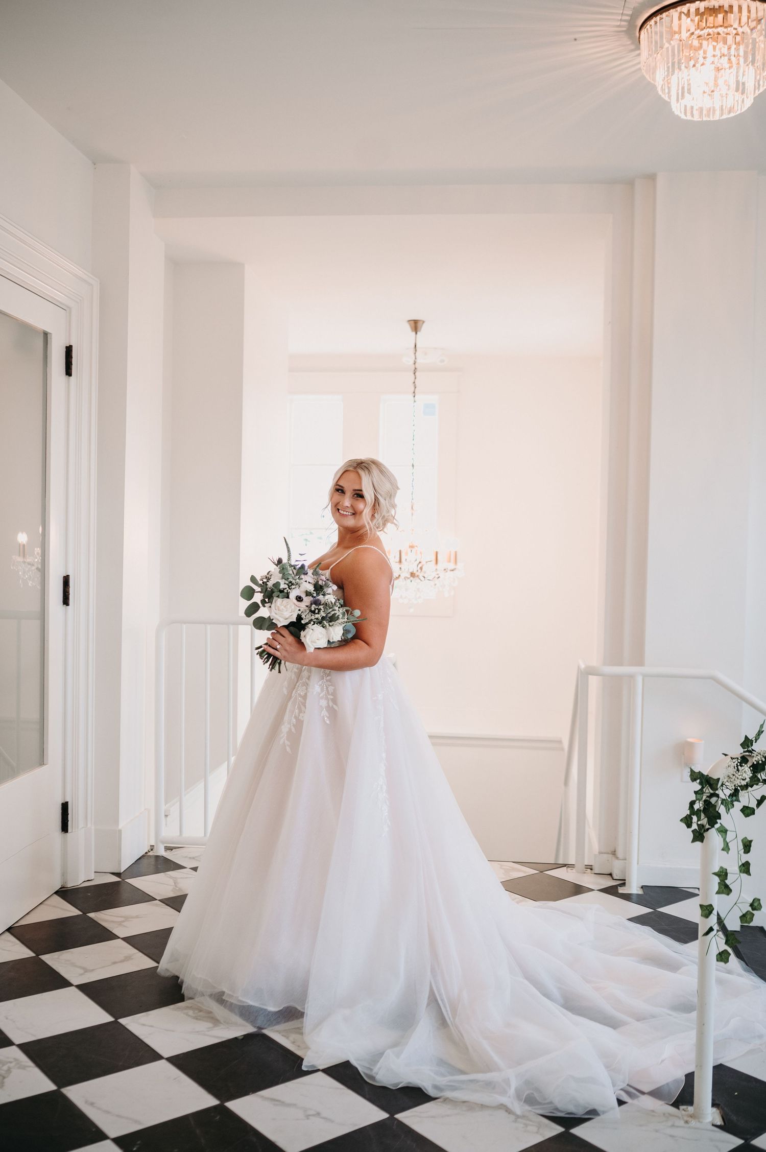 Bride holds bouquet while posing in stunning white gown against bright interior with crystal chandelier.