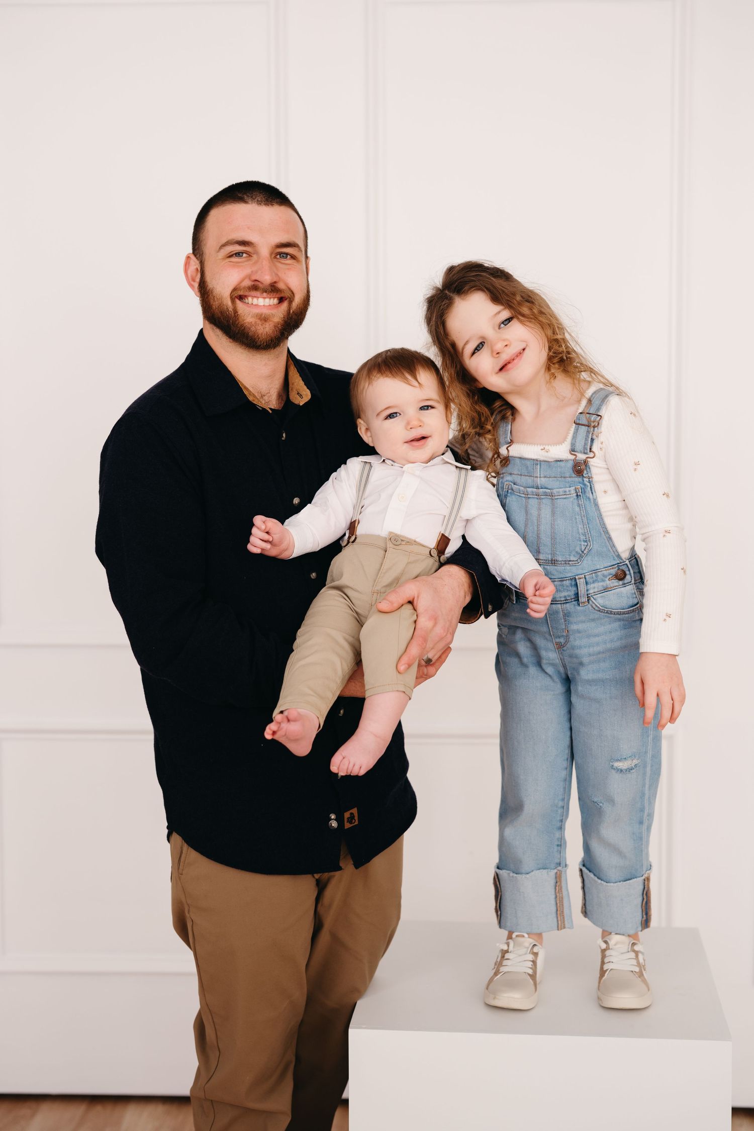 A family portrait against a white wall featuring a black-clad parent holding a baby and a child in denim overalls.