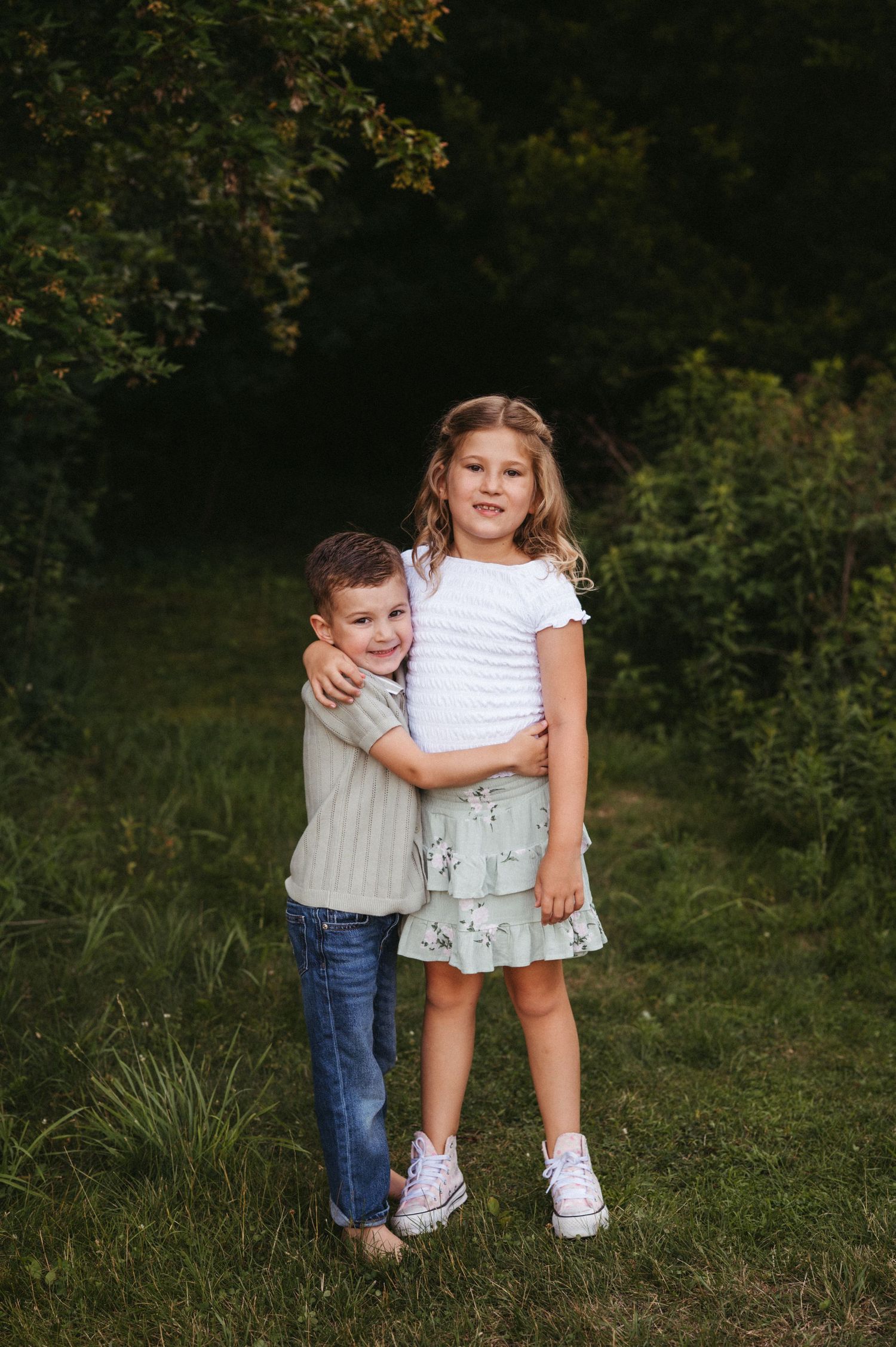 Two children embracing and smiling outdoors on a grassy area with trees and greenery in the background.