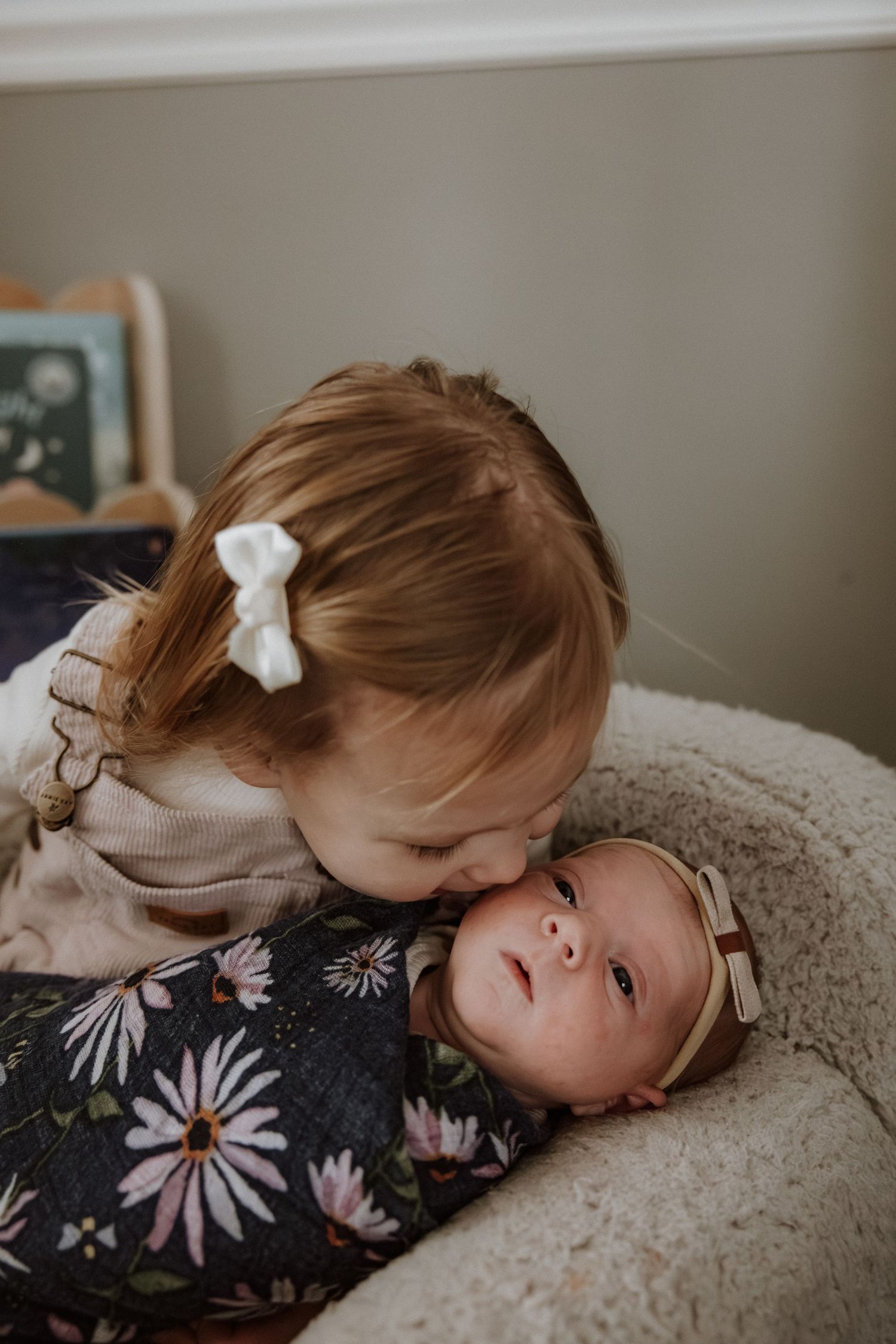 Two young children share a tender moment on a cozy blanket with one wearing a floral dress and white hair bow.