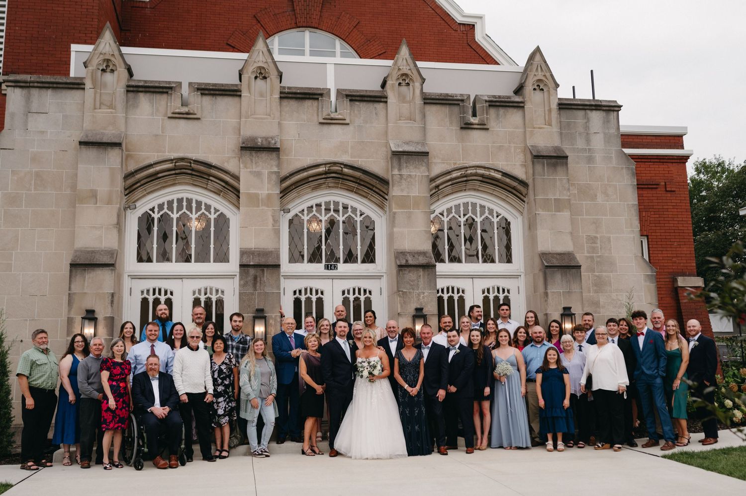 Large wedding party group photo with guests in front of stone church architecture.