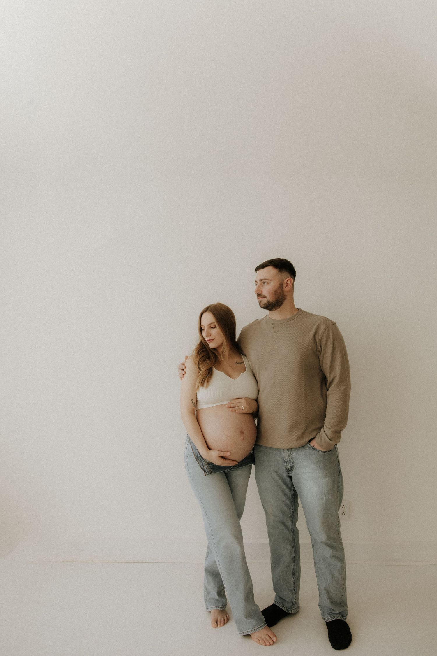Minimalist maternity photo of a couple in casual clothing standing against a white wall.