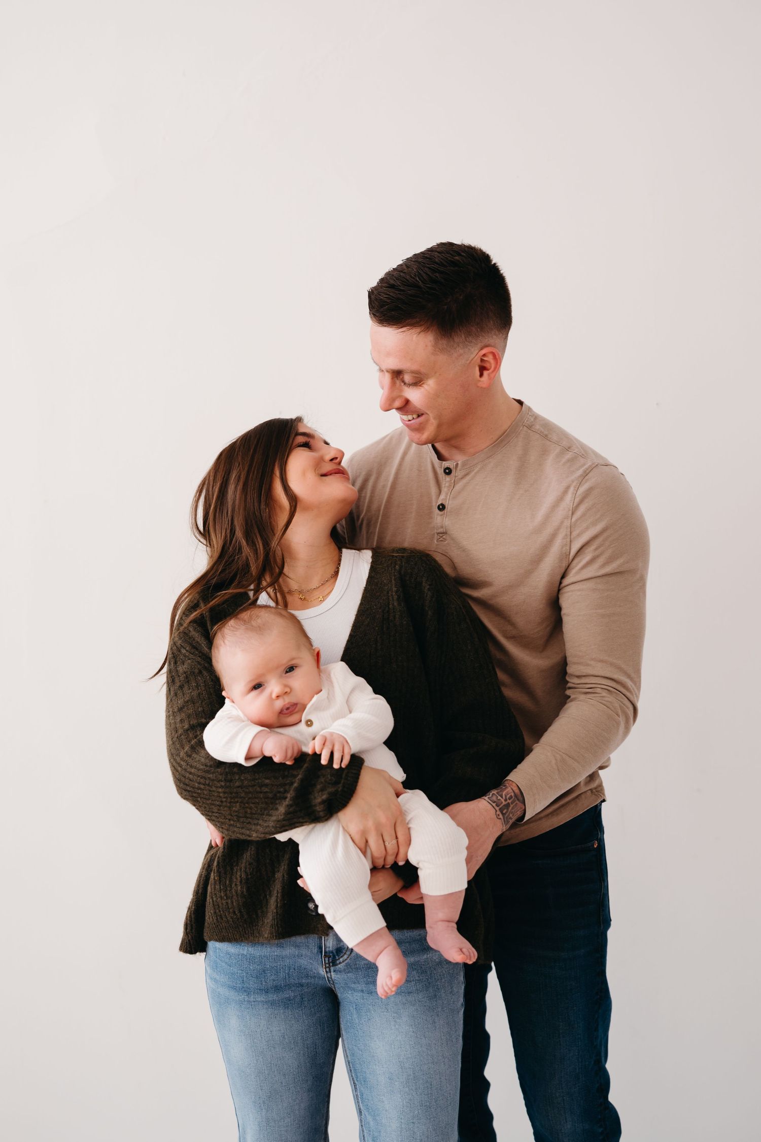 A couple in casual attire holds their baby against a light background in an intimate family portrait.