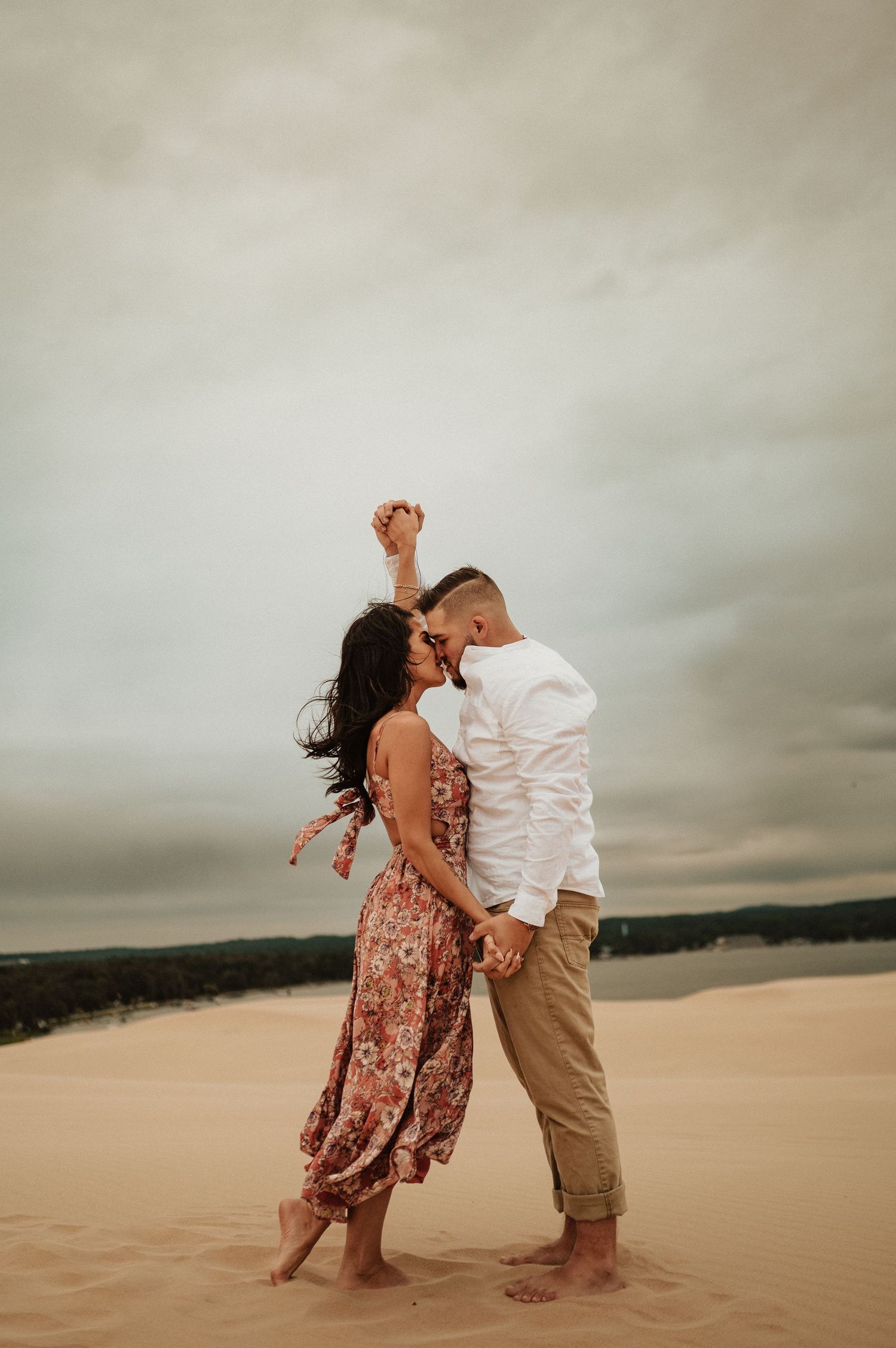 A couple shares an intimate moment while dancing on desert sand dunes under cloudy skies.