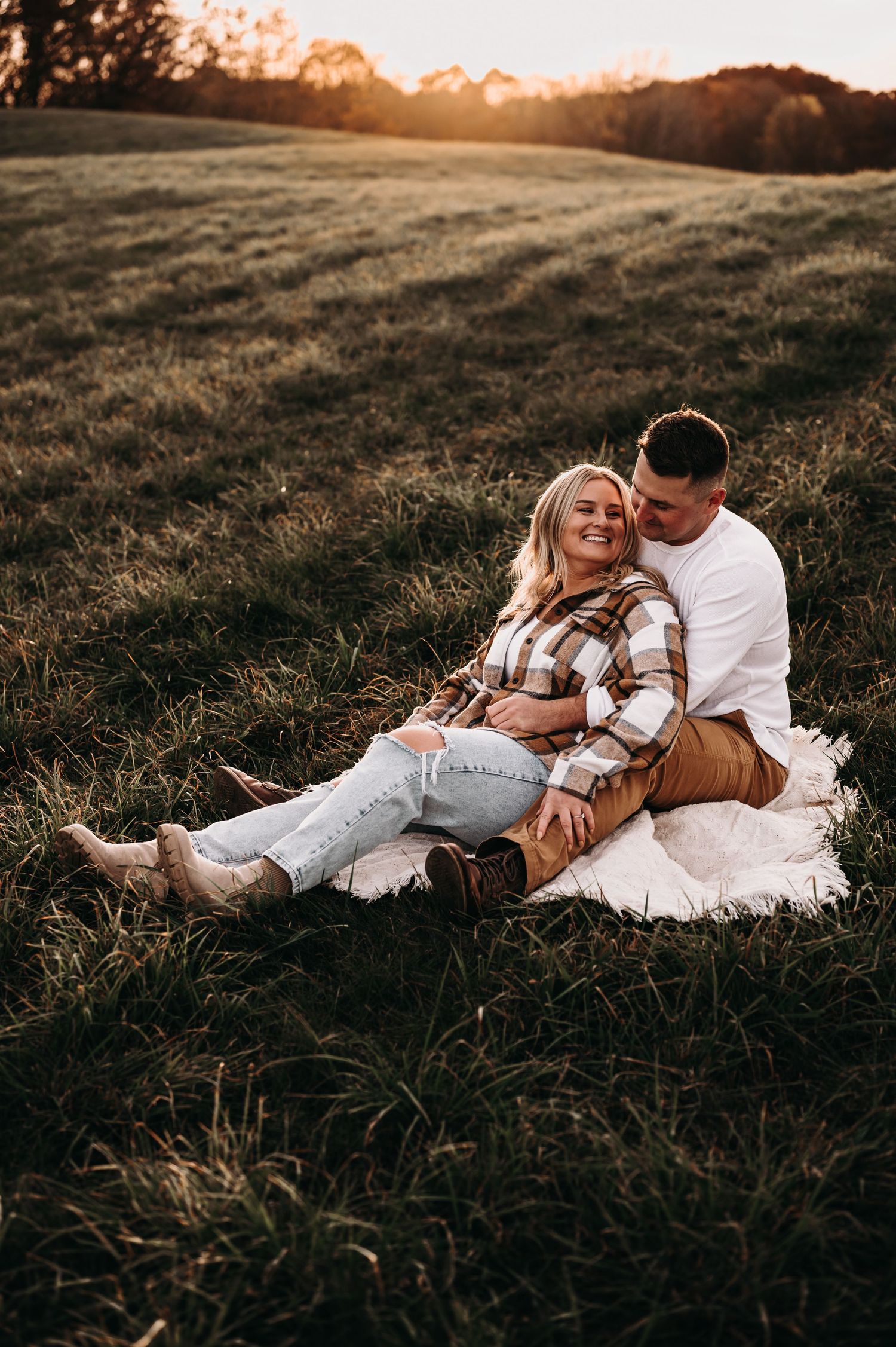 Romantic couple sitting on blanket in field at sunset wearing casual fall clothing.