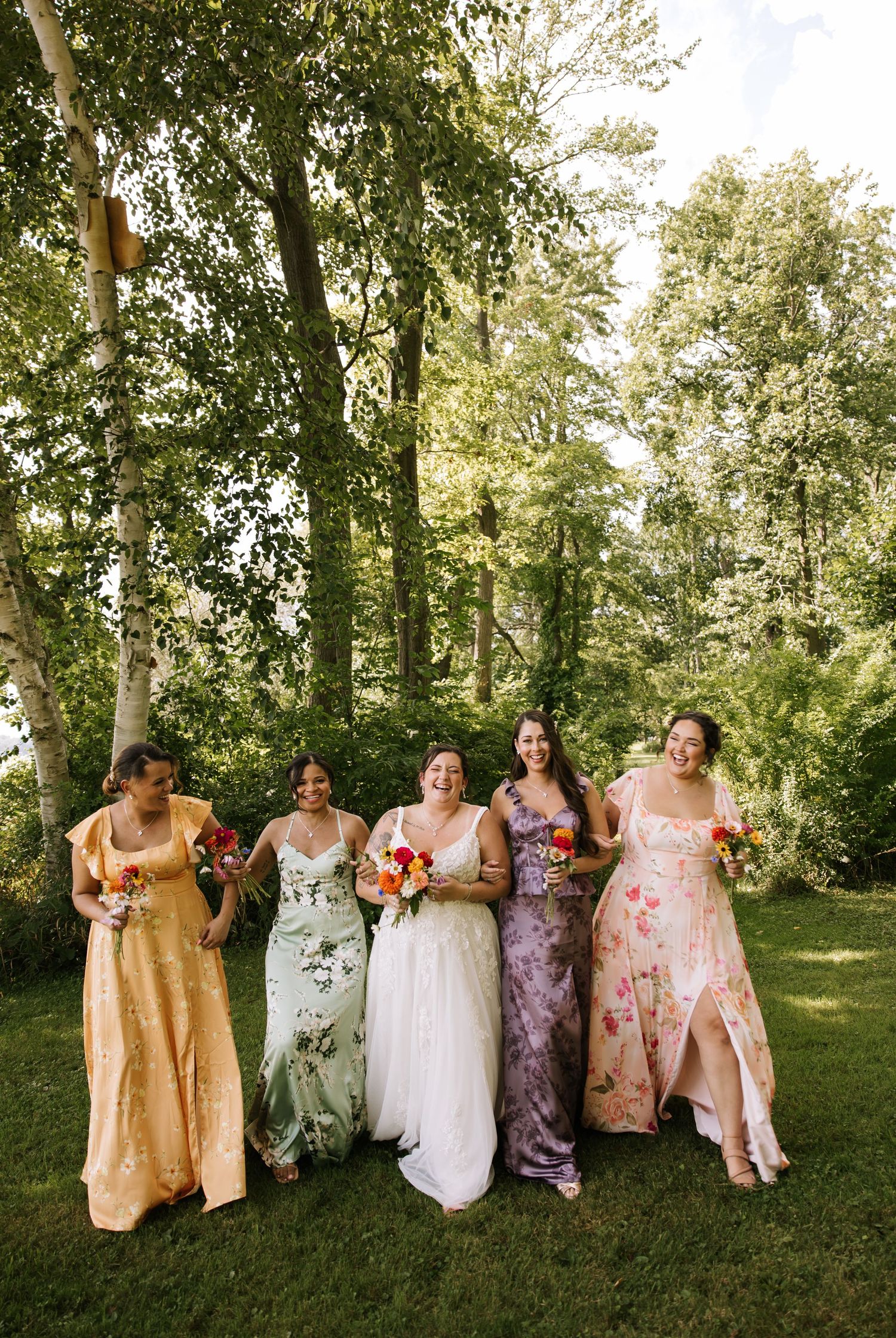 Wedding party in colorful floral dresses sitting on chairs arranged on a grassy lawn under trees.