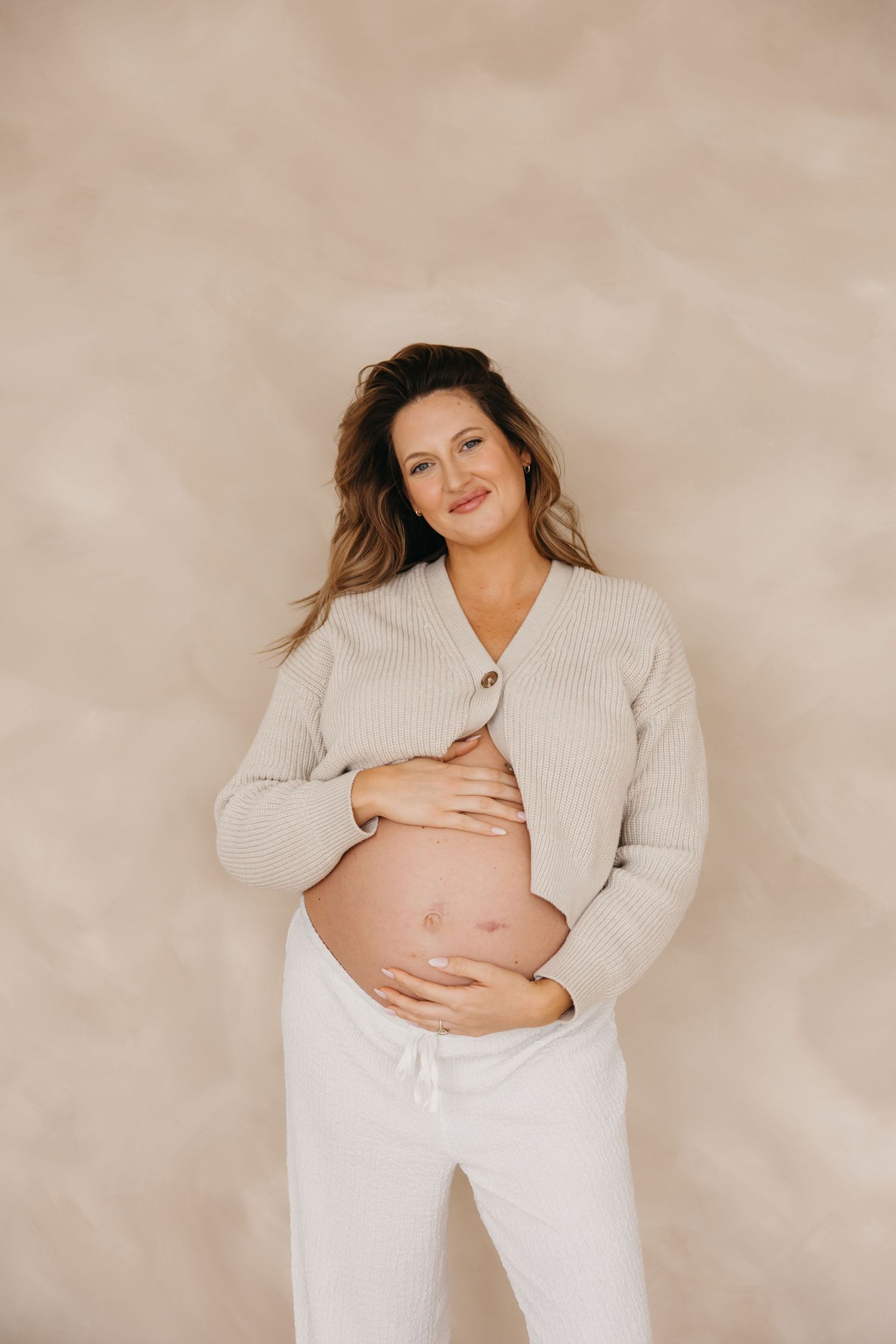 Smiling pregnant woman in beige ribbed cardigan and white pants lovingly holds her bare baby bump against beige wall.